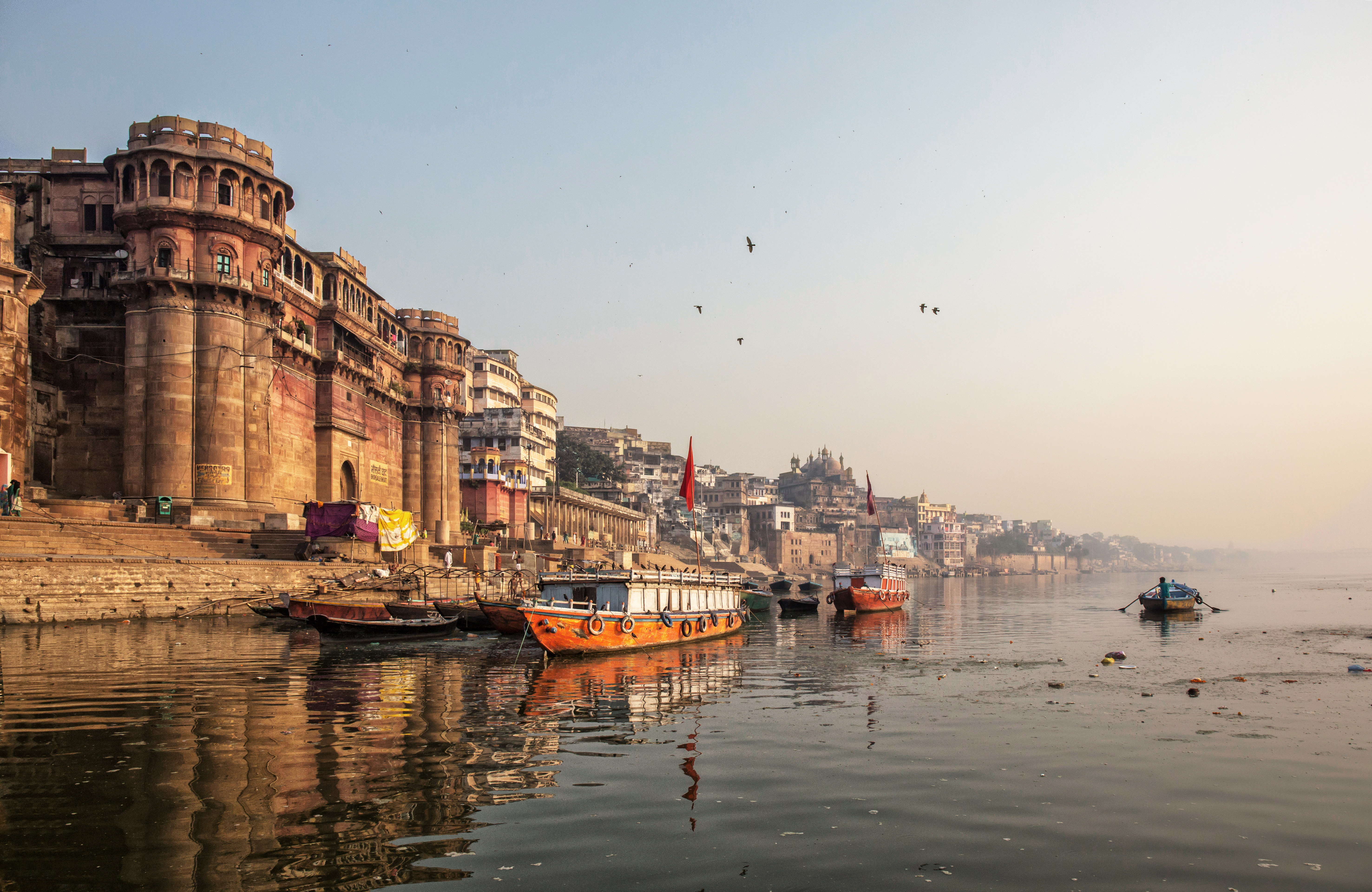 Boats float on a calm river beside stone ghats and buildings as birds circle in hazy sunrise light above.