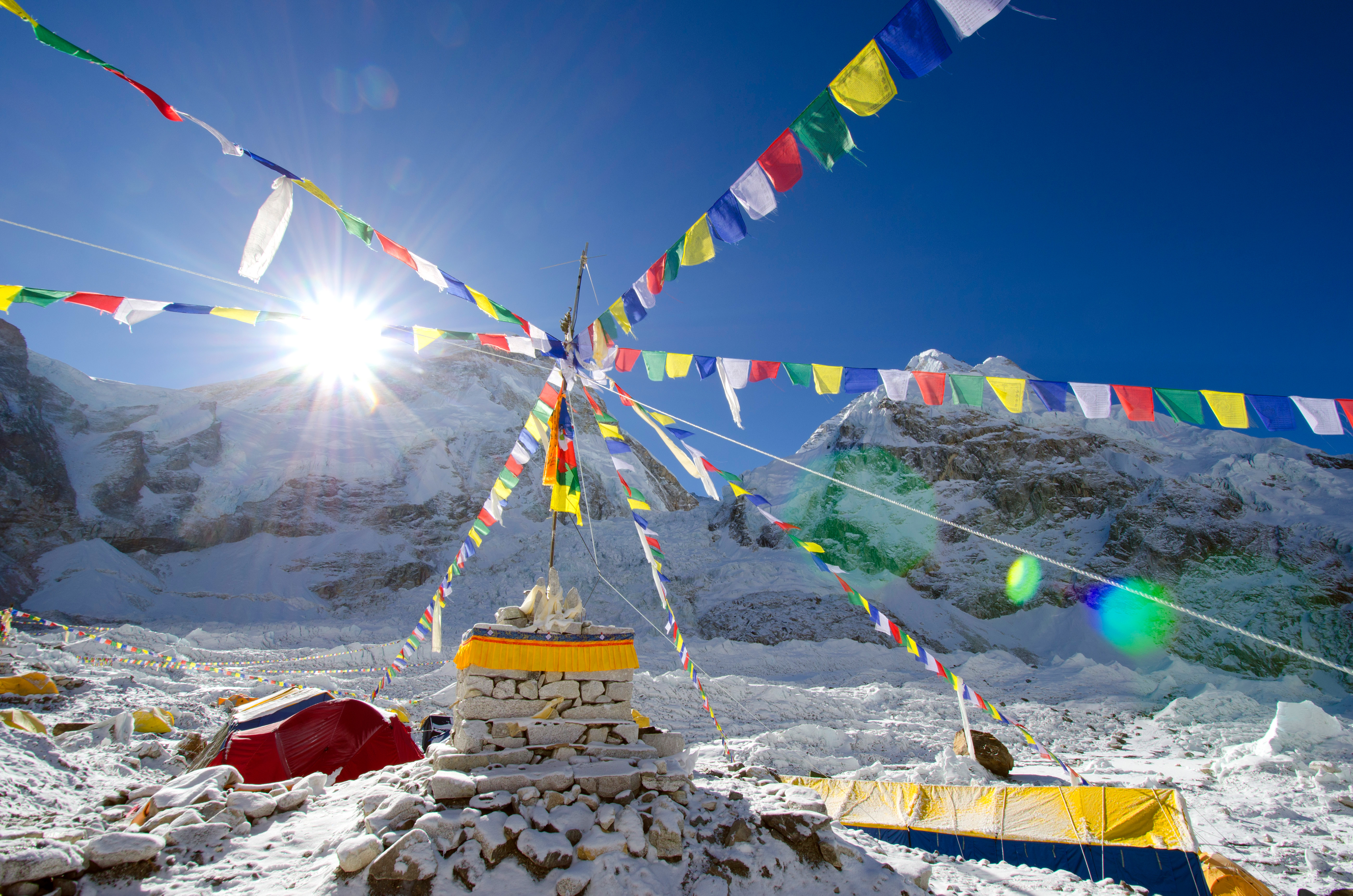 Colorful prayer flags radiate from a stupa as bright sun flares over snowy mountain peaks and windblown ice.