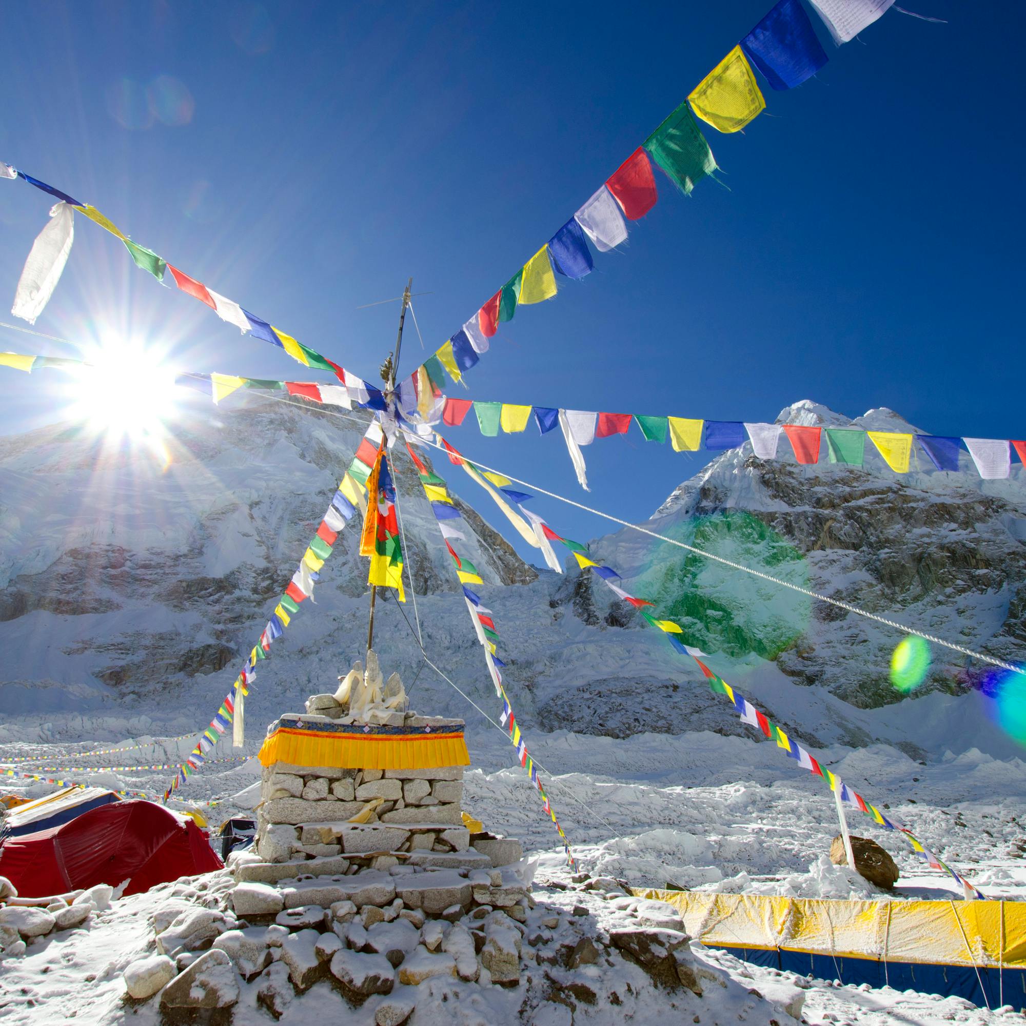 Colorful prayer flags radiate from a stupa as bright sun flares over snowy mountain peaks and windblown ice.