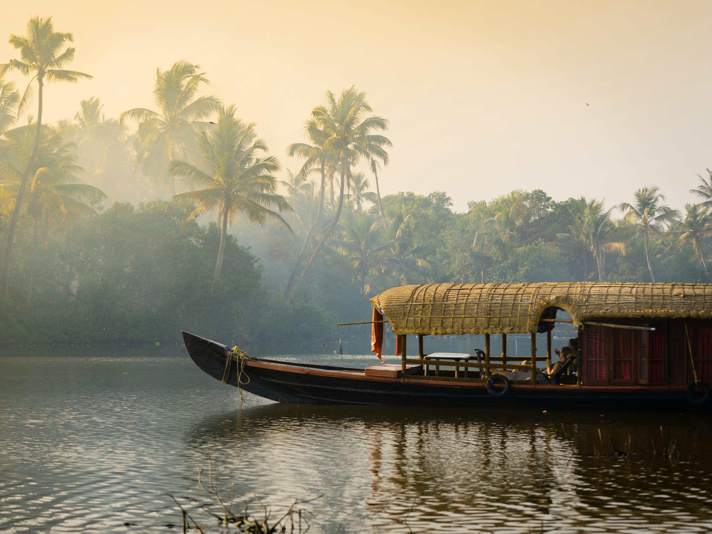 A houseboat glides along quiet backwaters lined with palms, with misty light and reflections on the rippled surface.