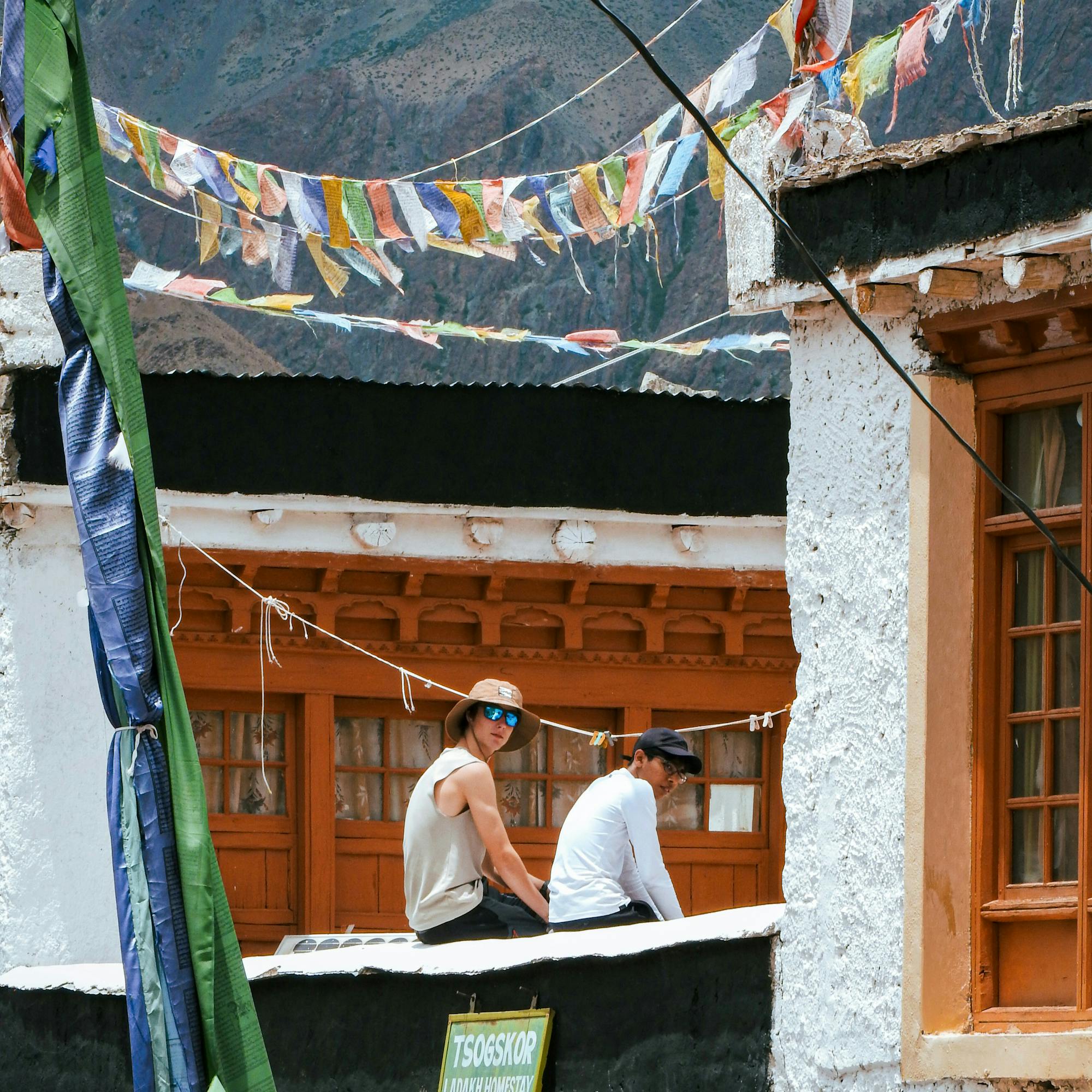 People walk through a monastery courtyard with prayer flags, whitewashed walls, and wooden windows under clear sky.