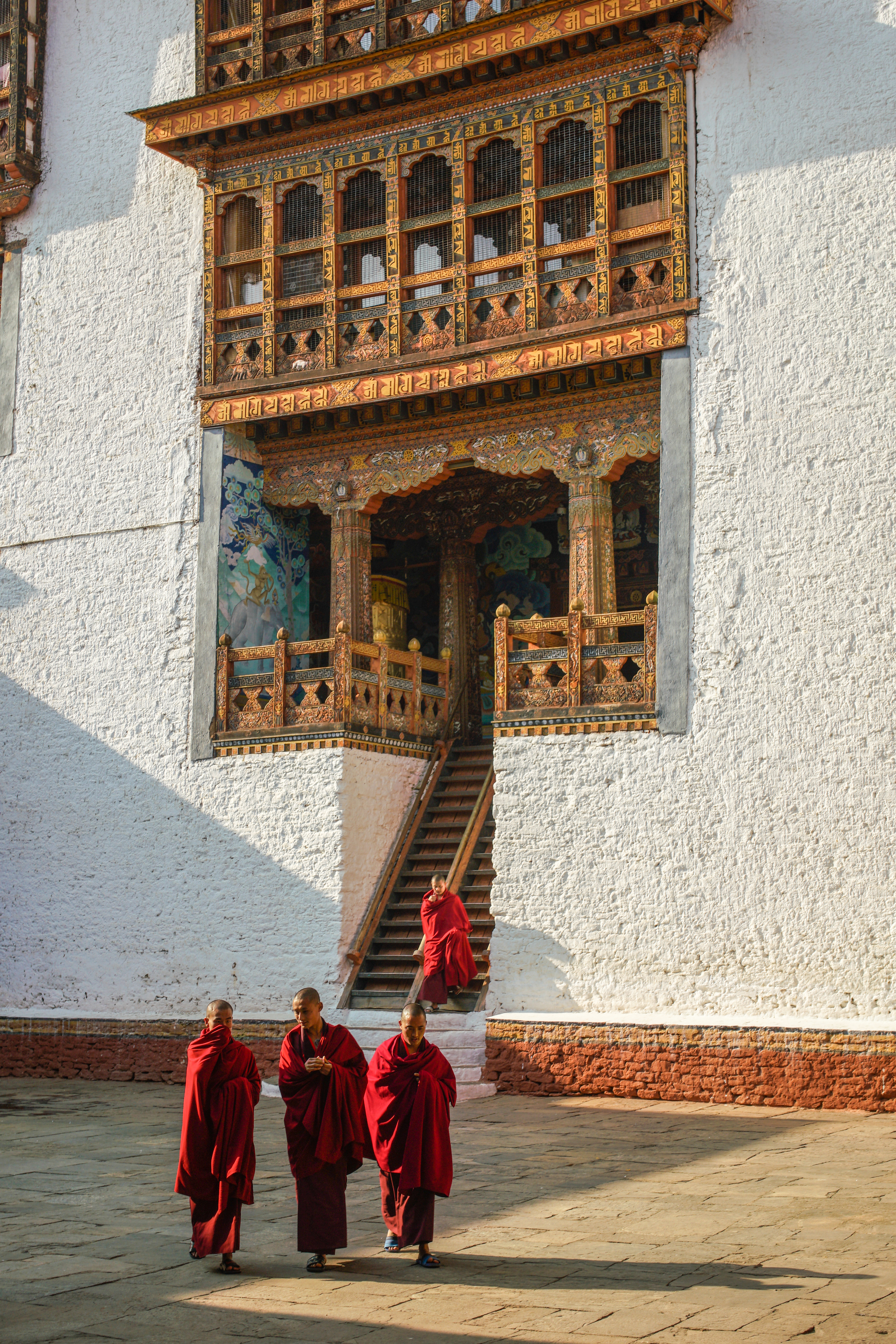 Monks in red robes walk beside a whitewashed building with ornate windows, casting shadows on a stone courtyard.