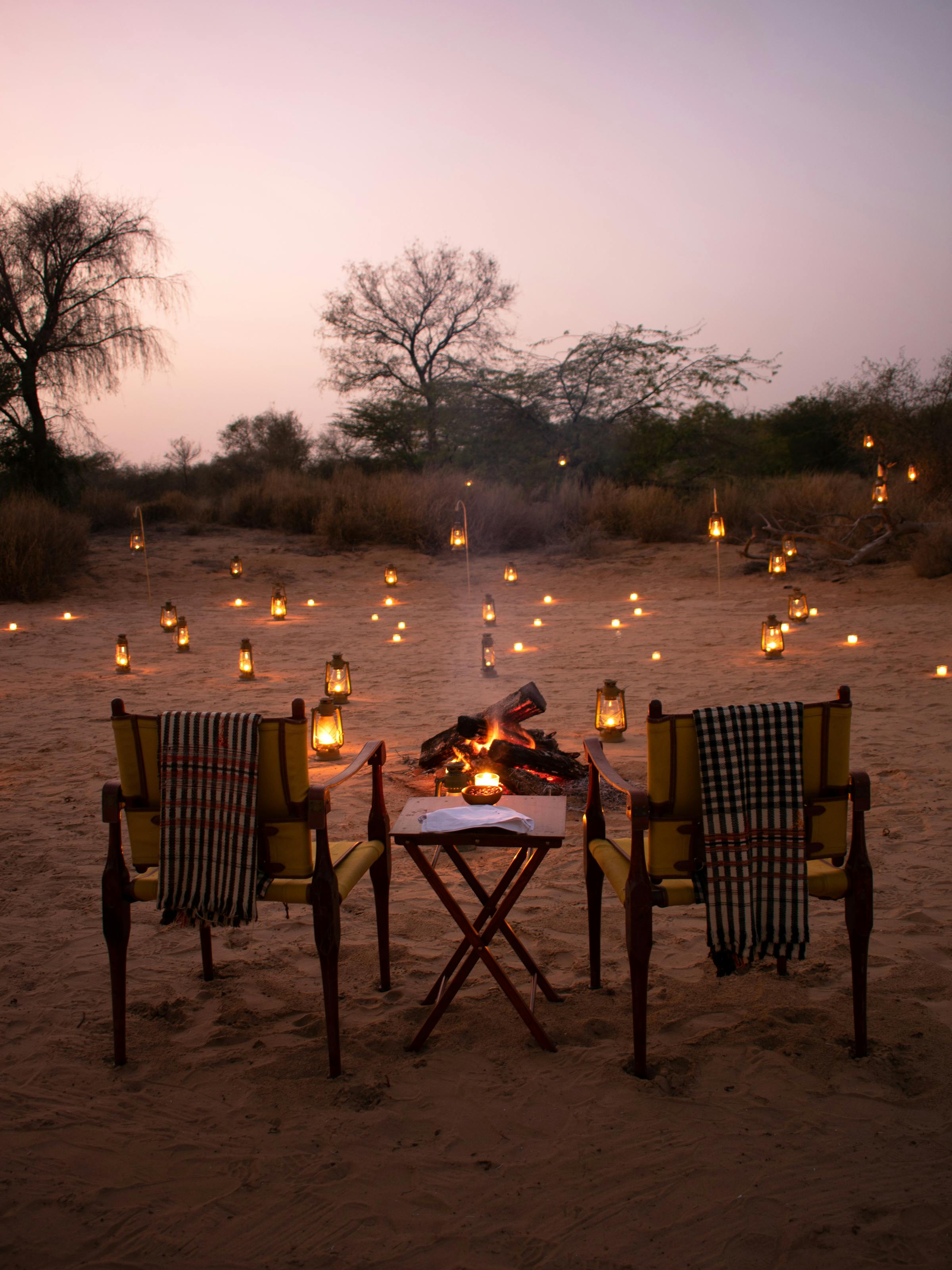 A candlelit table set for two sits on sandy ground, surrounded by lanterns and empty chairs at twilight glow.
