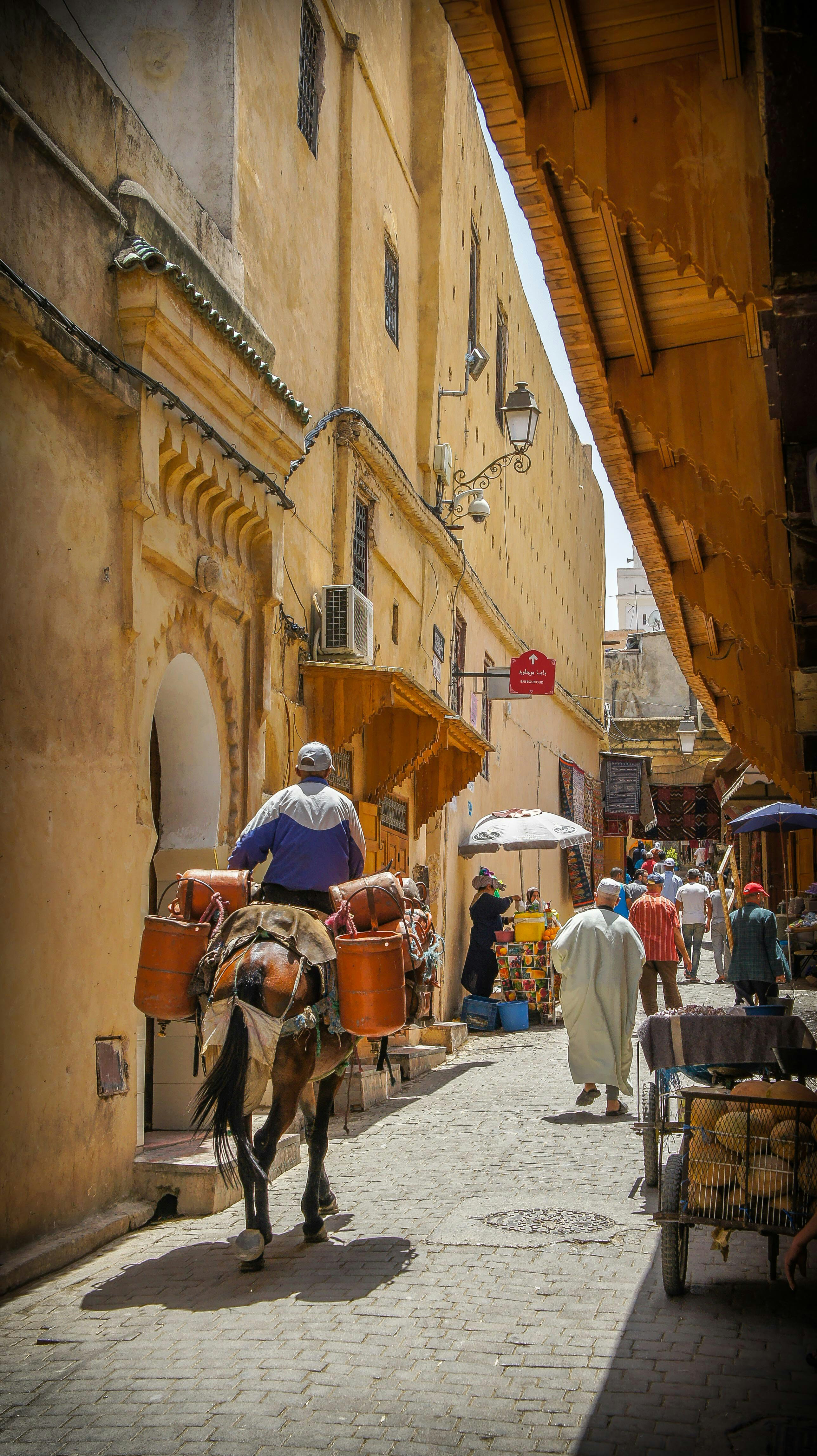 Narrow alleyway lined with yellow walls as a man on a horse rides past pedestrians, with sunlight cutting through above.
