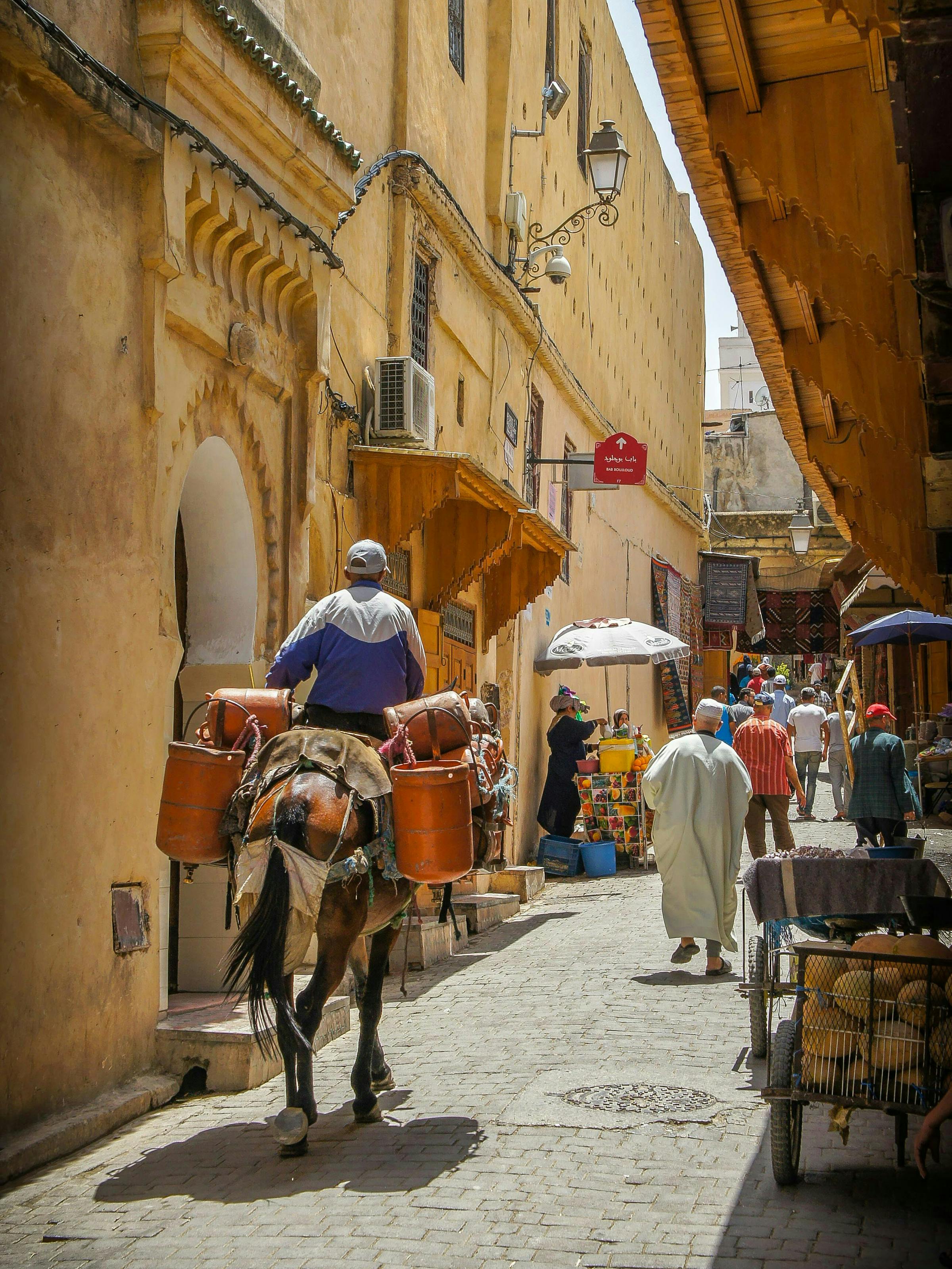 Narrow alleyway lined with yellow walls as a man on a horse rides past pedestrians, with sunlight cutting through above.