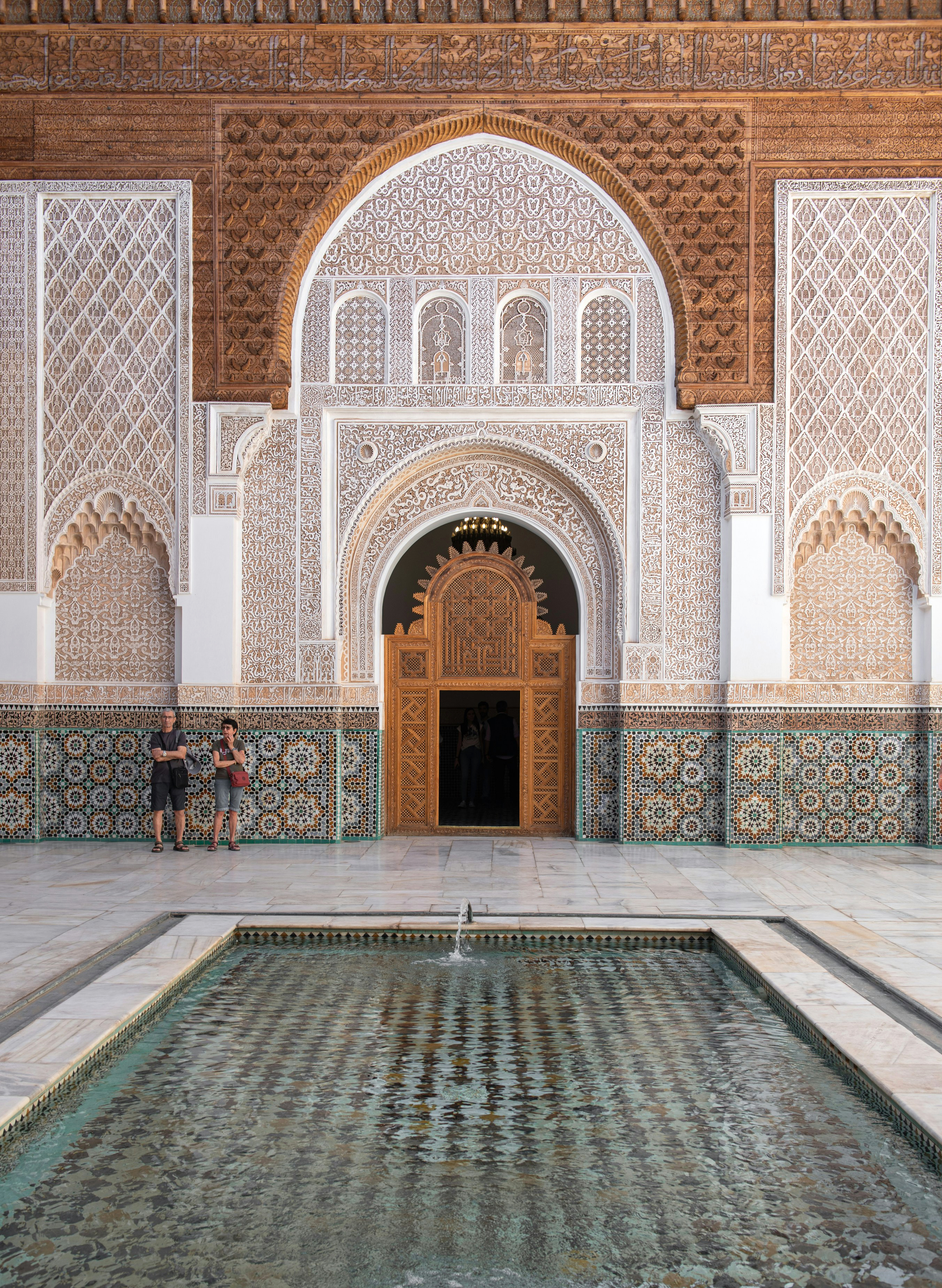 Ornate arched doorway with carved plaster and mosaic tiles opens onto a quiet courtyard with patterned stone floor.
