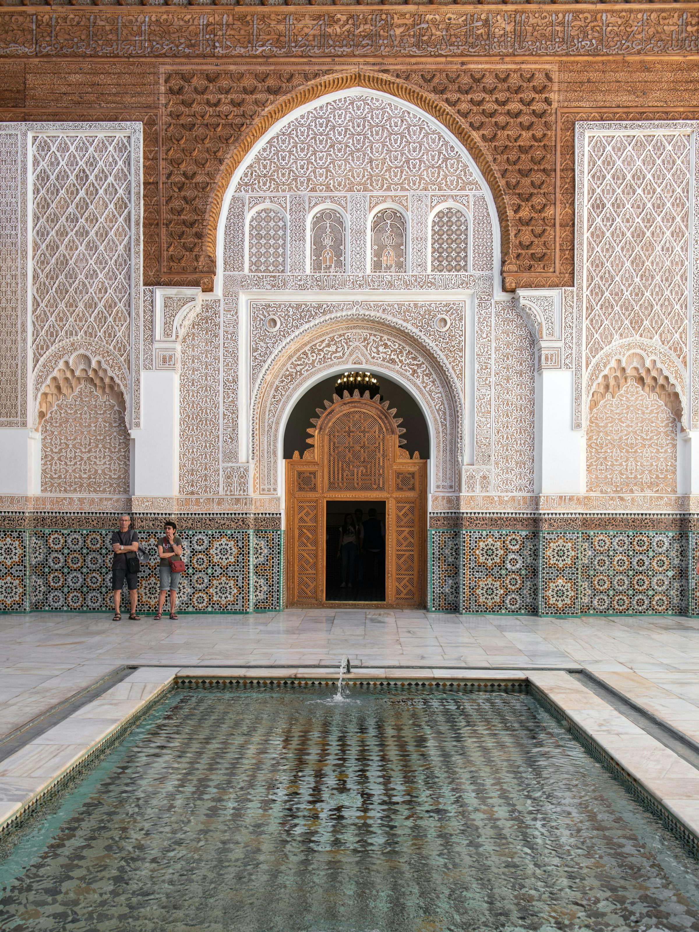 Ornate arched doorway with carved plaster and mosaic tiles opens onto a quiet courtyard with patterned stone floor.