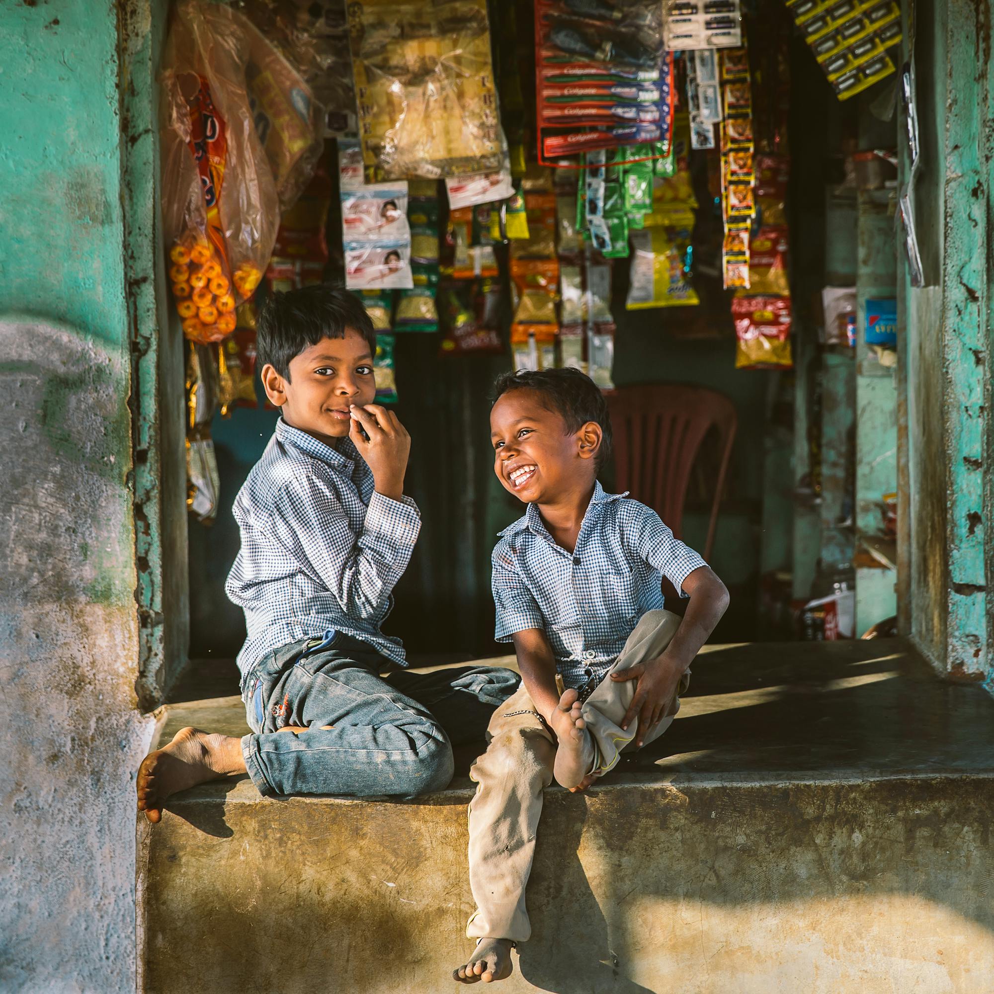Two boys sit at a small shop entrance, chatting and smiling amid stacked goods and a weathered green wall.