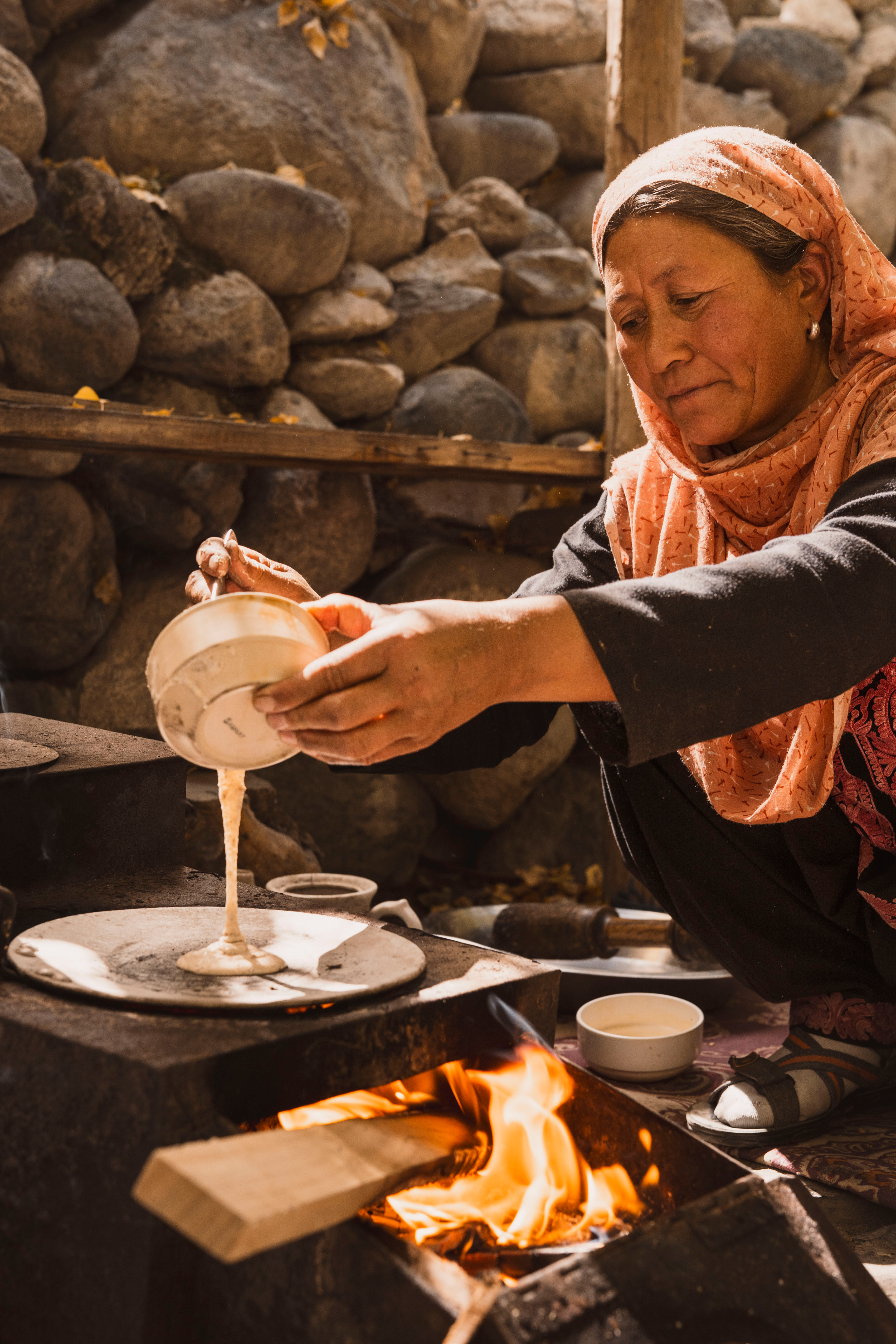 A woman in a headscarf pours tea beside an open fire, with metal pots and a stone wall forming the rustic kitchen.