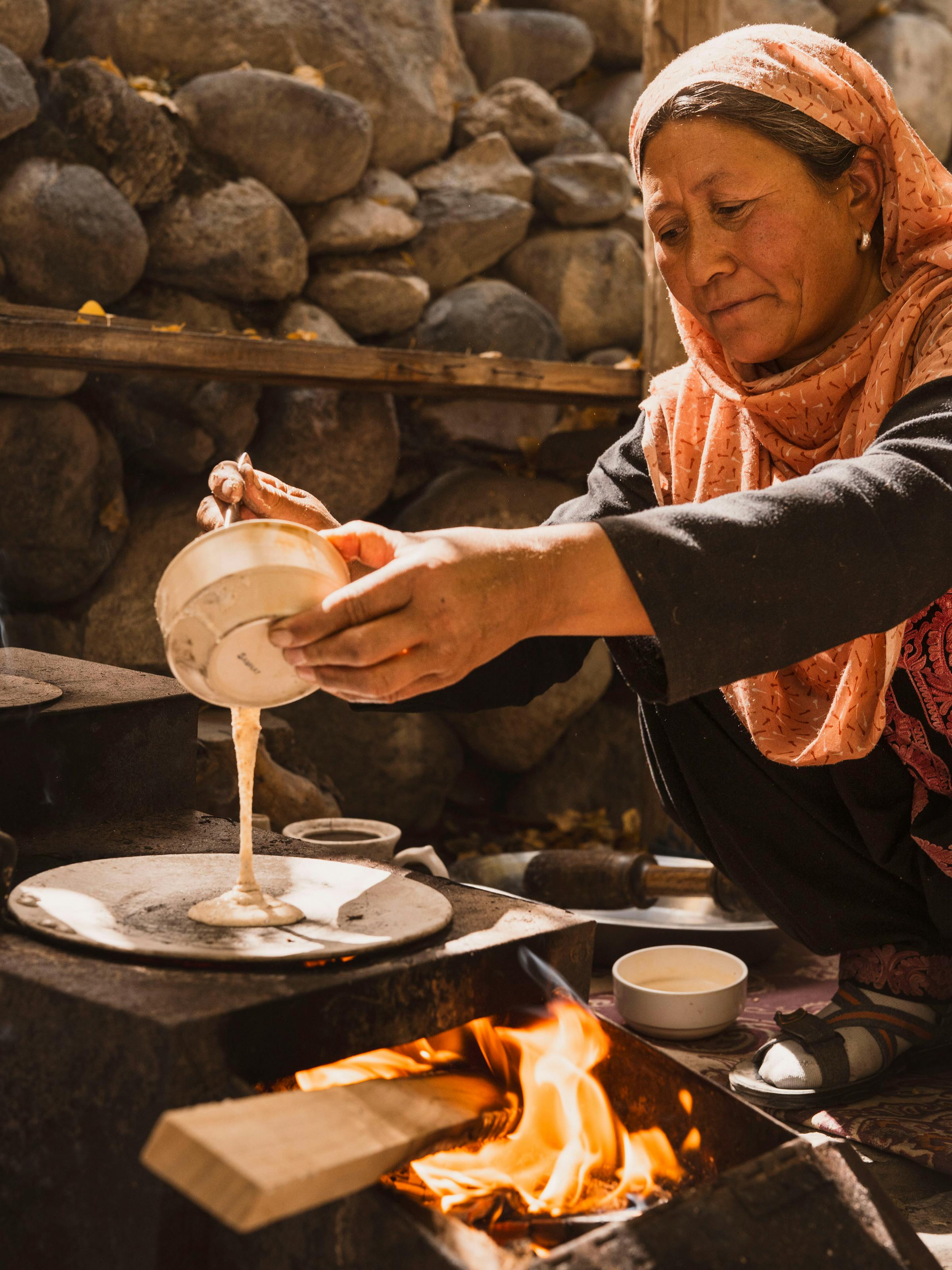 A woman in a headscarf pours tea beside an open fire, with metal pots and a stone wall forming the rustic kitchen.