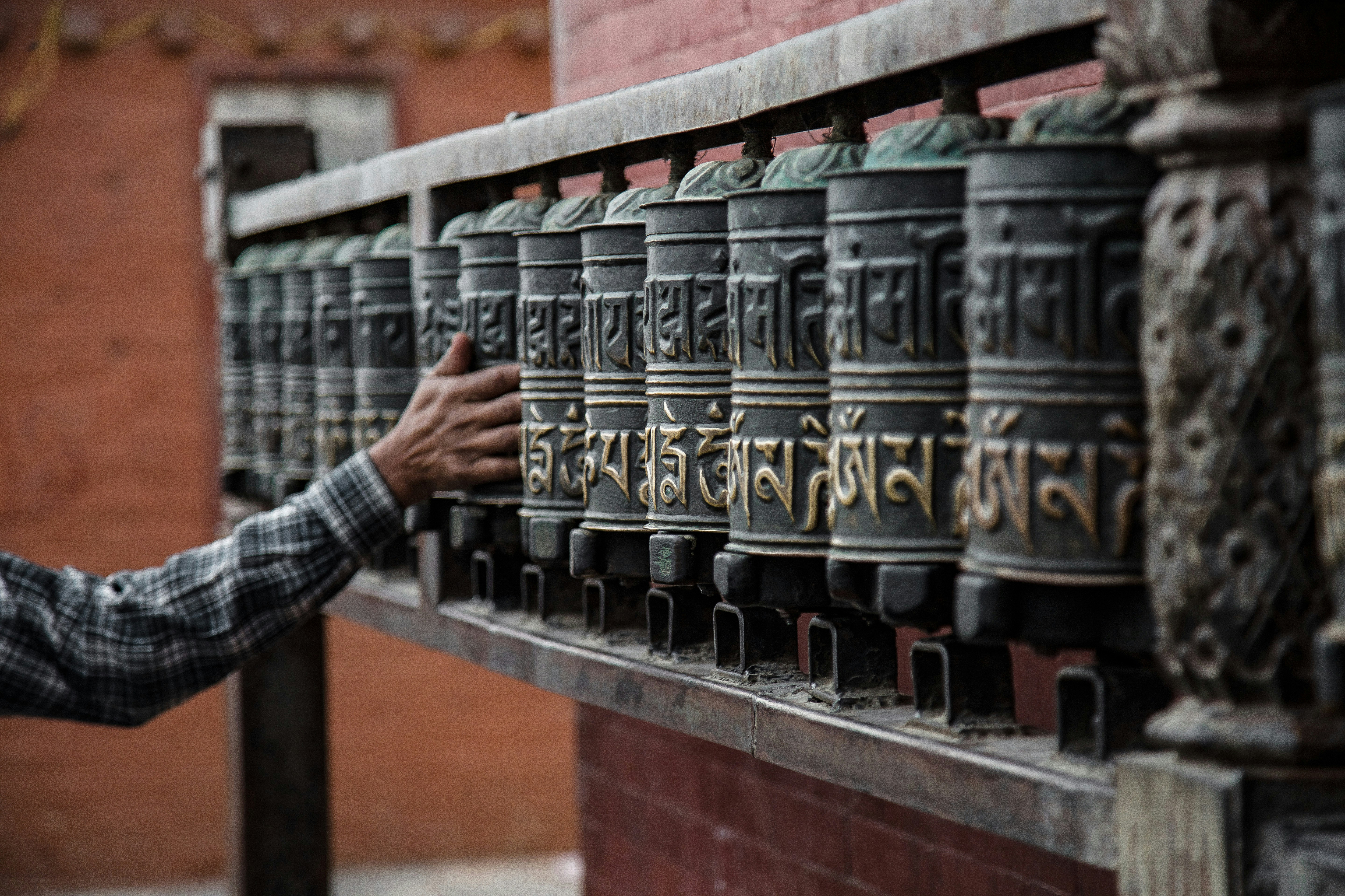 A hand spins a row of carved prayer wheels mounted on a wall, with warm metal surfaces catching the light.