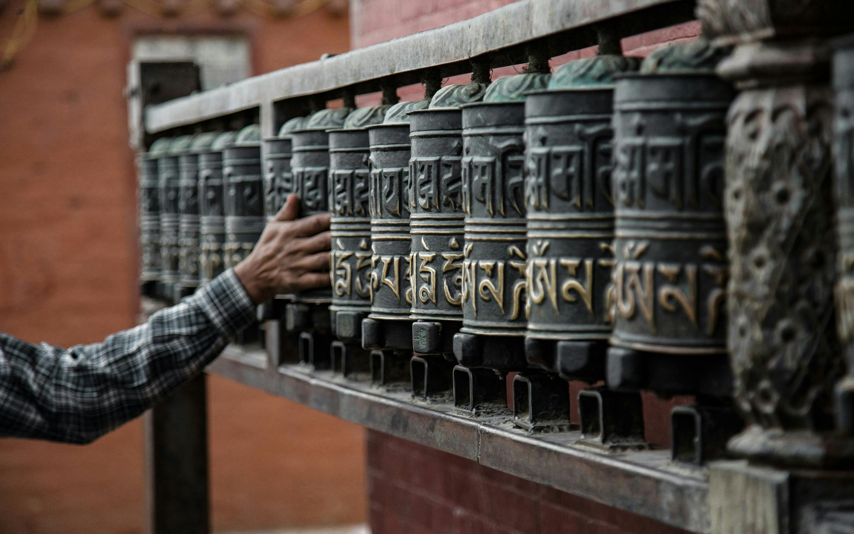 A hand spins a row of carved prayer wheels mounted on a wall, with warm metal surfaces catching the light.