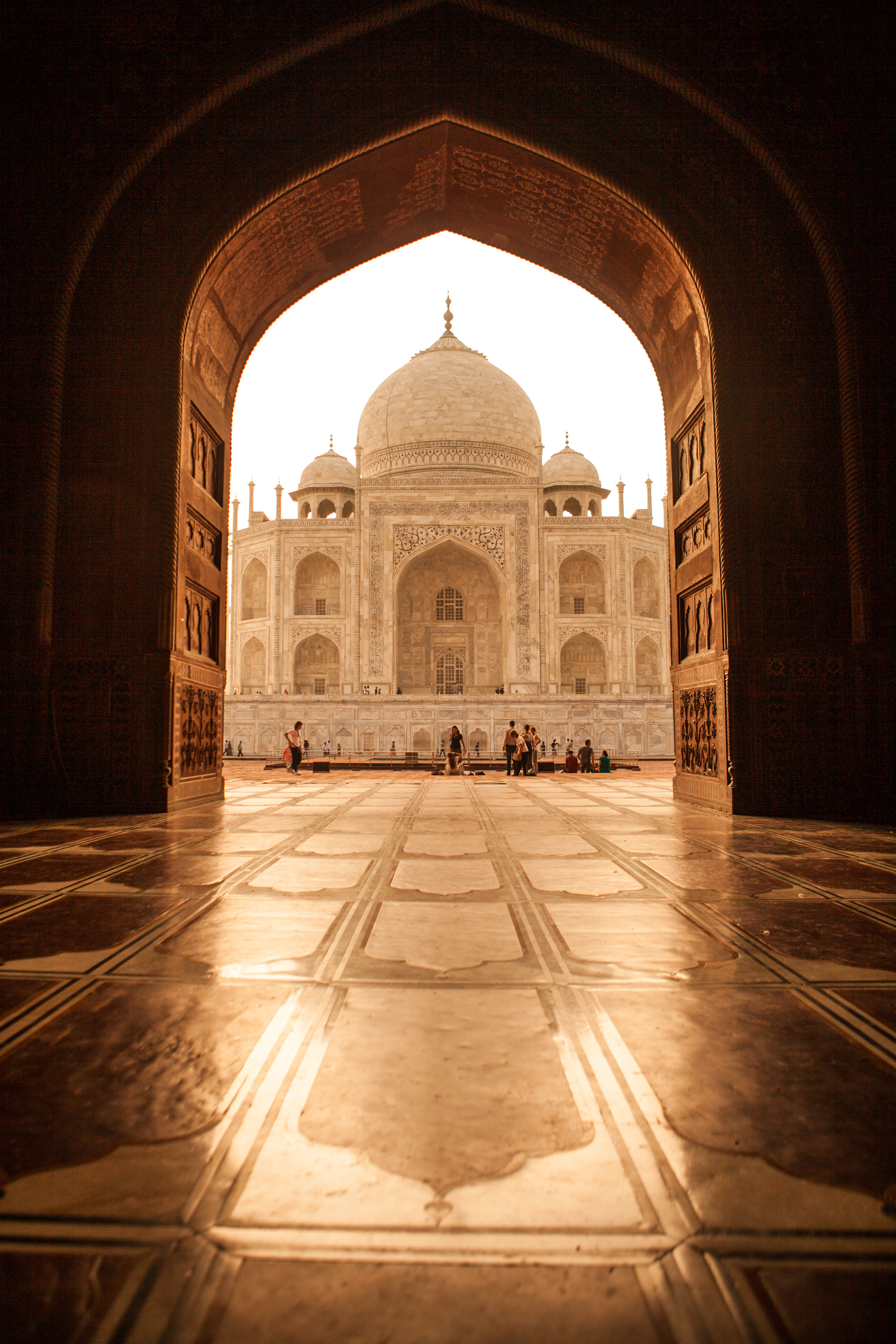 The Taj Mahal glows beyond a dark archway, with marble flooring leading to a bright courtyard in soft morning light.
