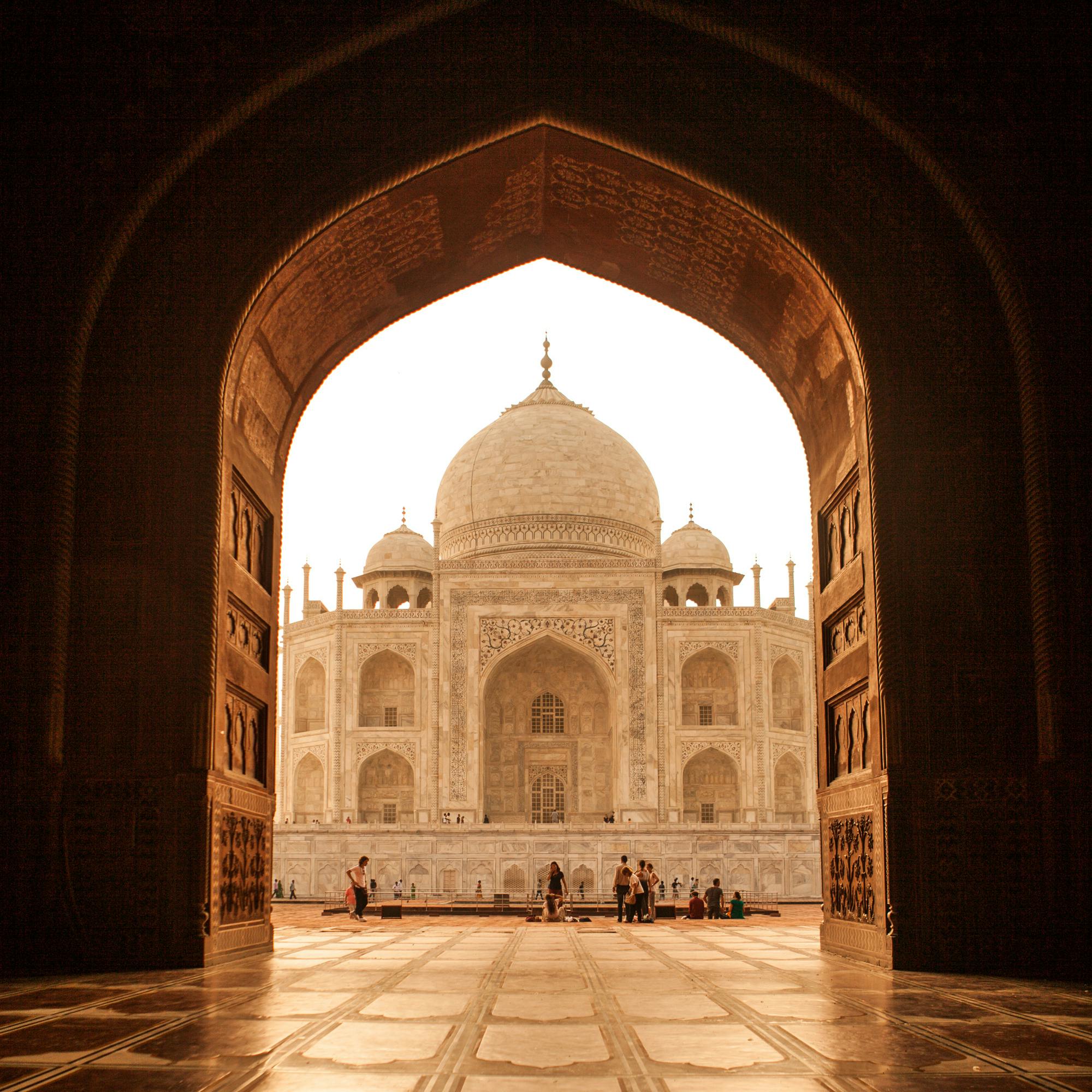 The Taj Mahal glows beyond a dark archway, with marble flooring leading to a bright courtyard in soft morning light.