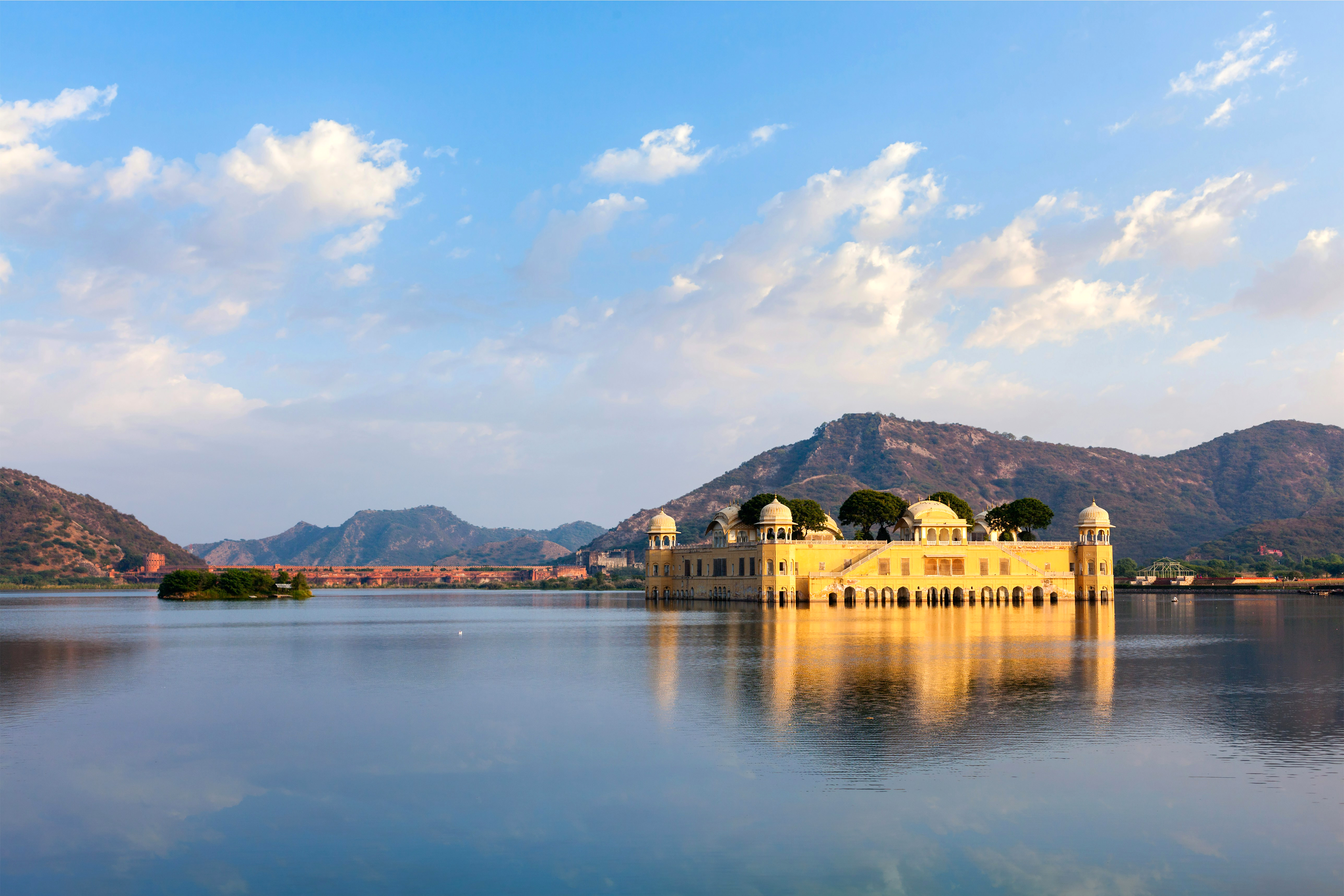 A pale palace sits in the middle of a lake, with blue hills and scattered clouds reflected across the still water.