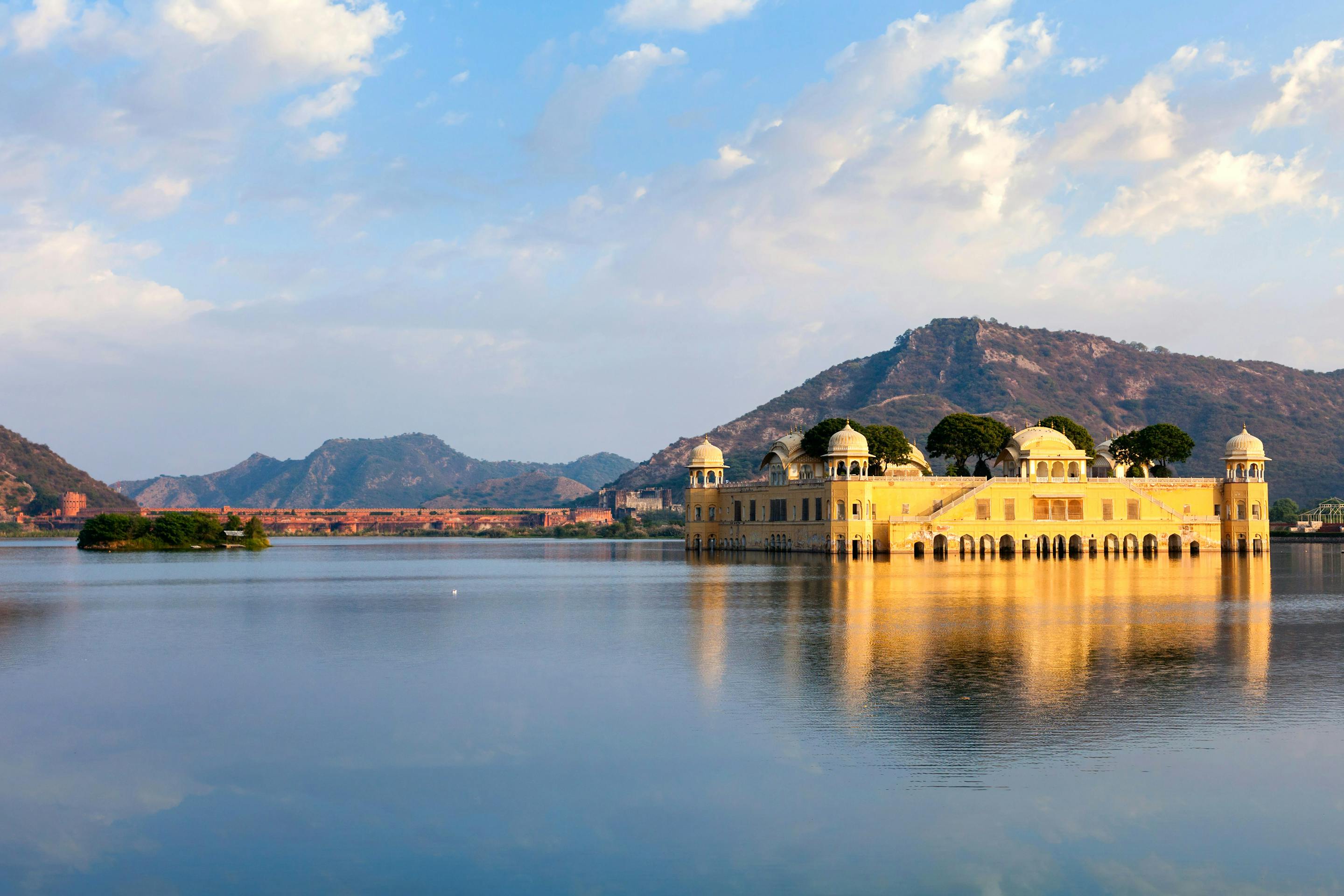 A pale palace sits in the middle of a lake, with blue hills and scattered clouds reflected across the still water.