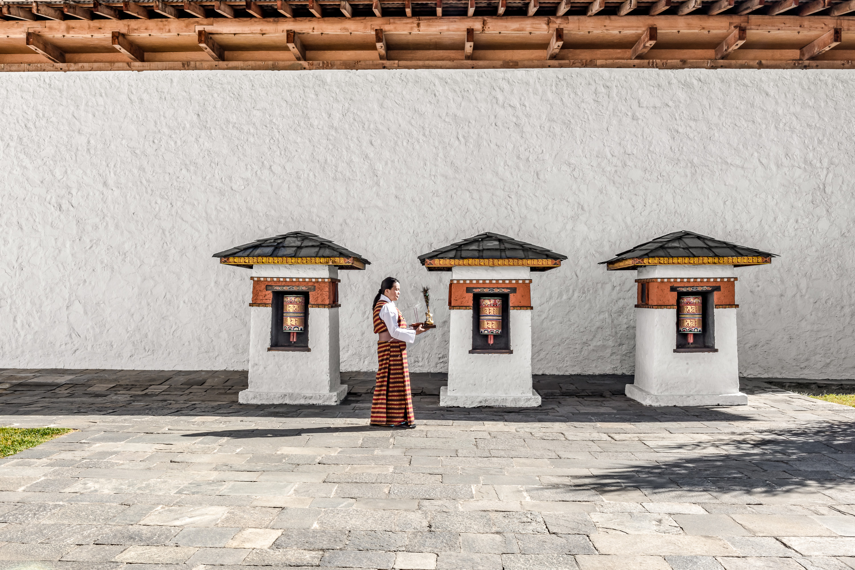 A person in traditional dress stands in a quiet walled courtyard, with simple windows and long shadows on stone.