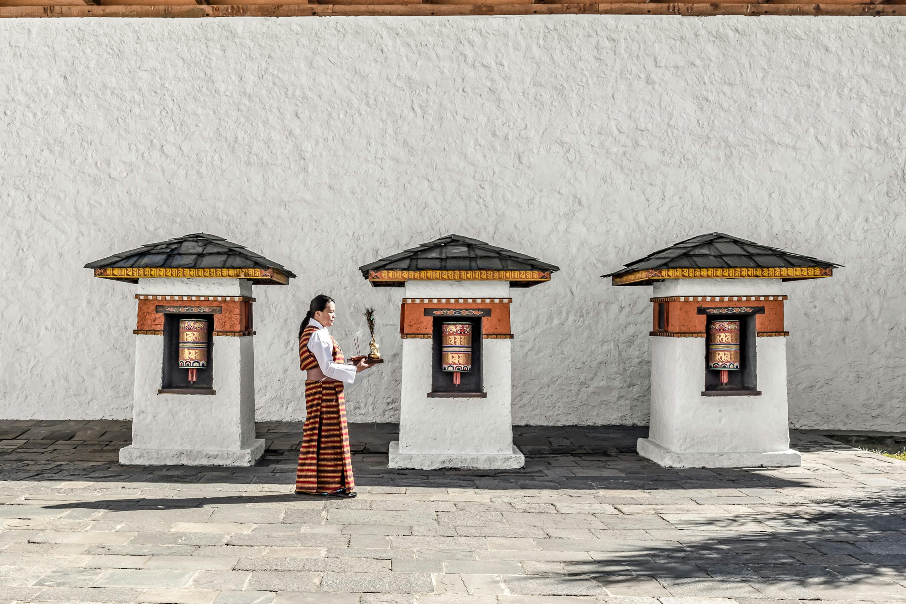 A person in traditional dress stands in a quiet walled courtyard, with simple windows and long shadows on stone.