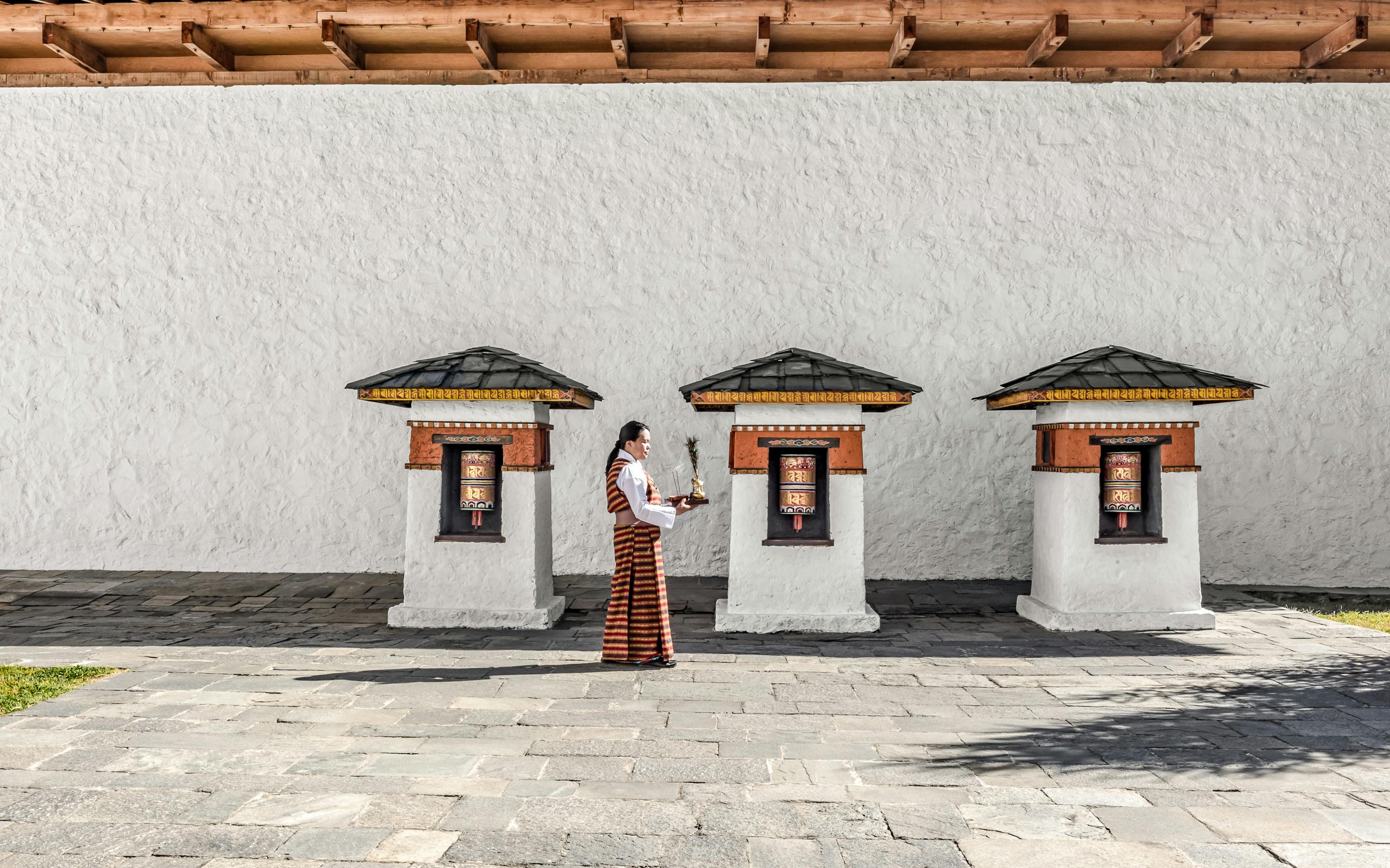 A person in traditional dress stands in a quiet walled courtyard, with simple windows and long shadows on stone.