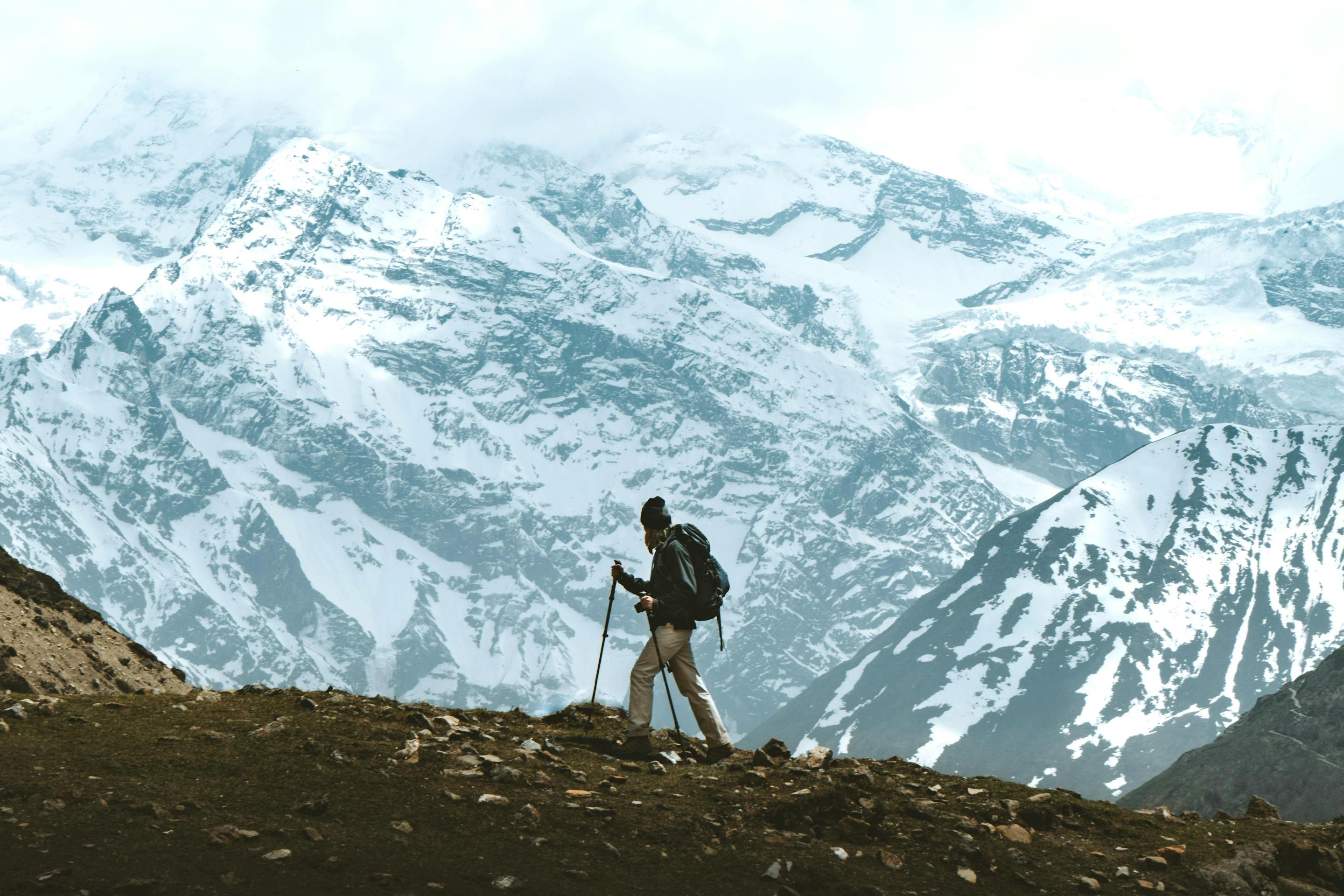A lone hiker walks along a dark ridge with vast snowcapped mountains and glaciers stretching across the horizon.
