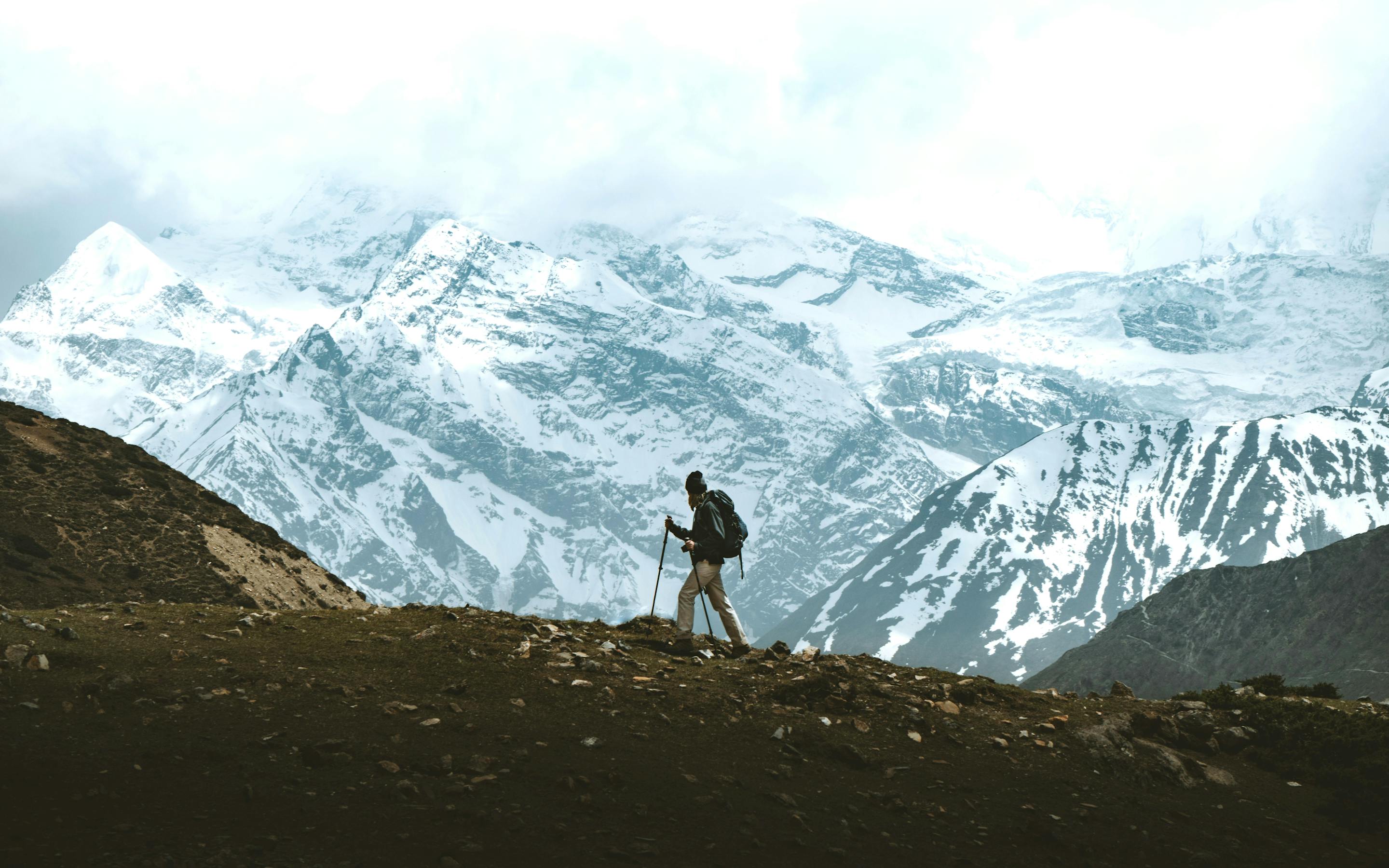 A lone hiker walks along a dark ridge with vast snowcapped mountains and glaciers stretching across the horizon.