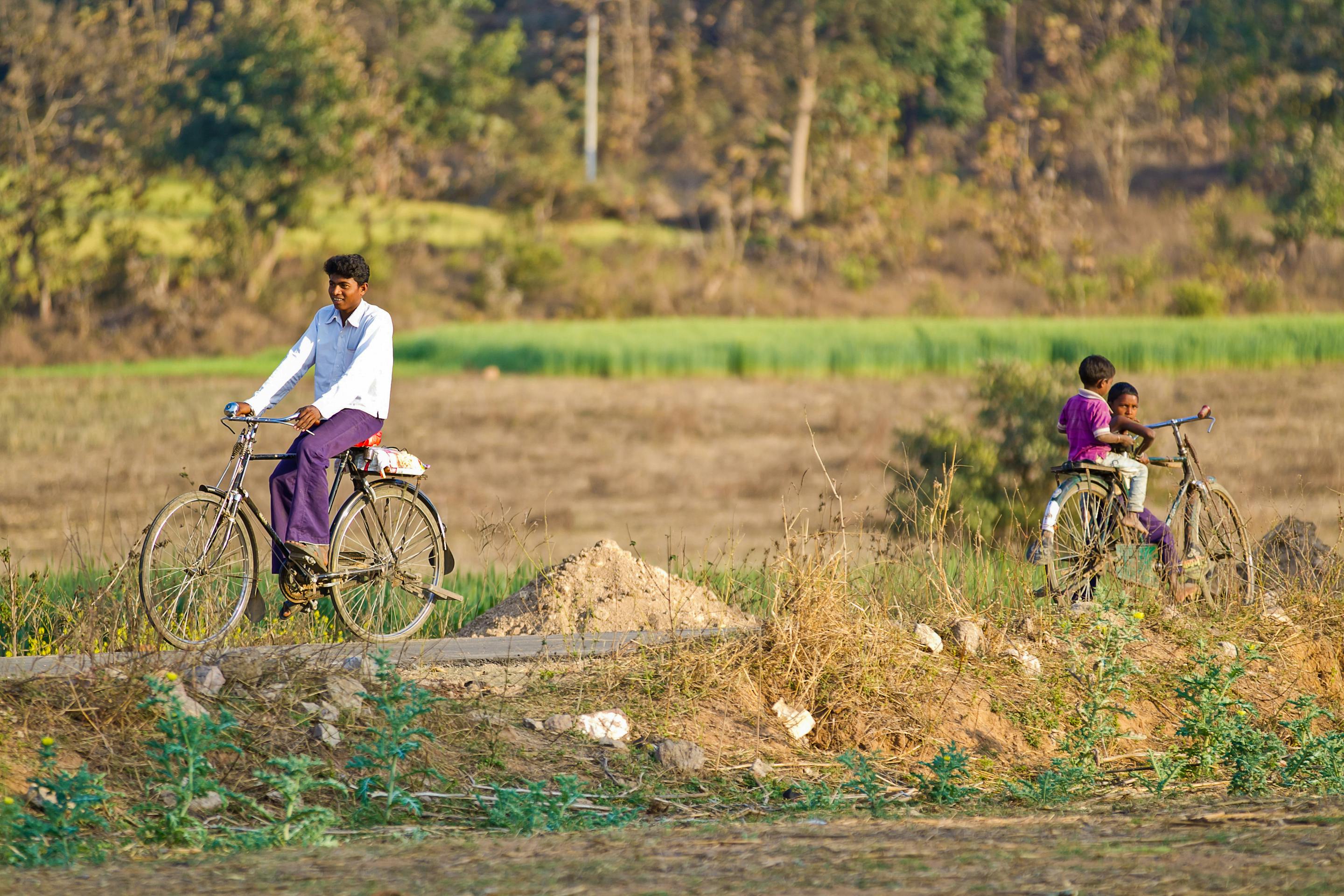 An adult and child ride bicycles along a dirt path through rural fields, with trees and low hills in the background.
