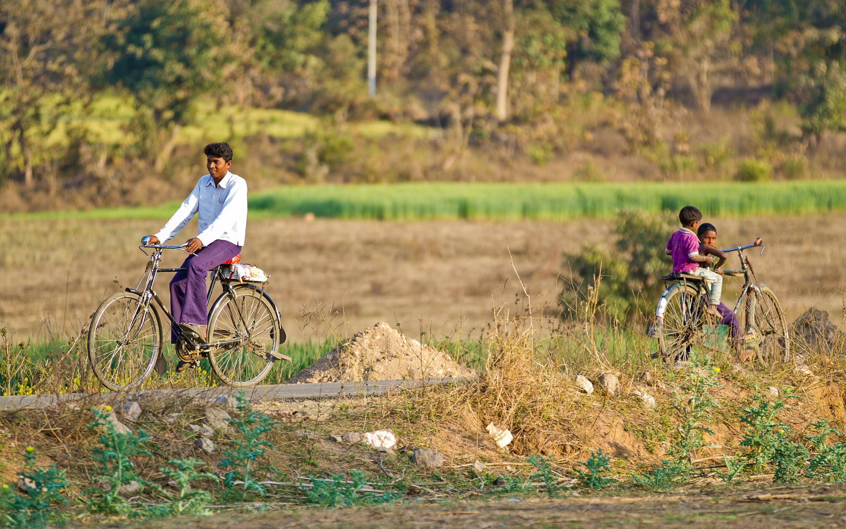 An adult and child ride bicycles along a dirt path through rural fields, with trees and low hills in the background.