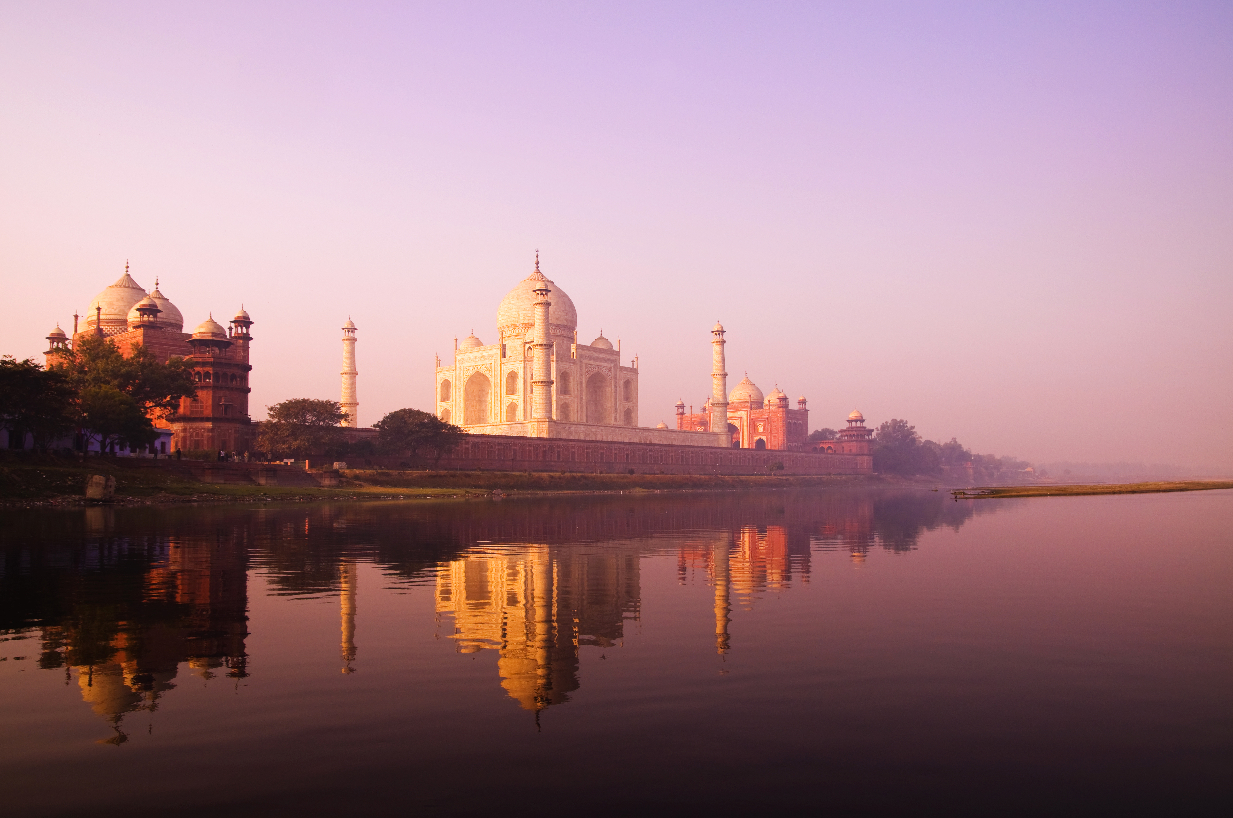 The Taj Mahal reflects in a calm pool at dawn, with pastel sky and dark silhouettes of nearby buildings framing it.