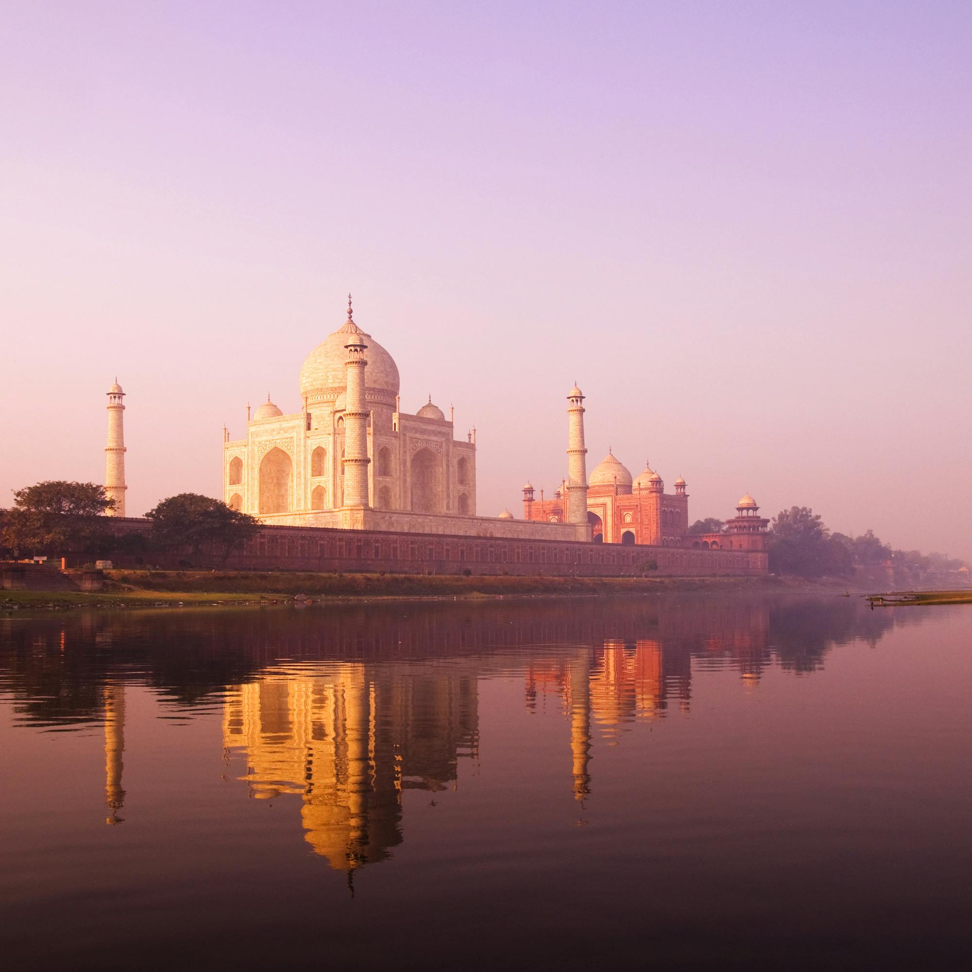 The Taj Mahal reflects in a calm pool at dawn, with pastel sky and dark silhouettes of nearby buildings framing it.