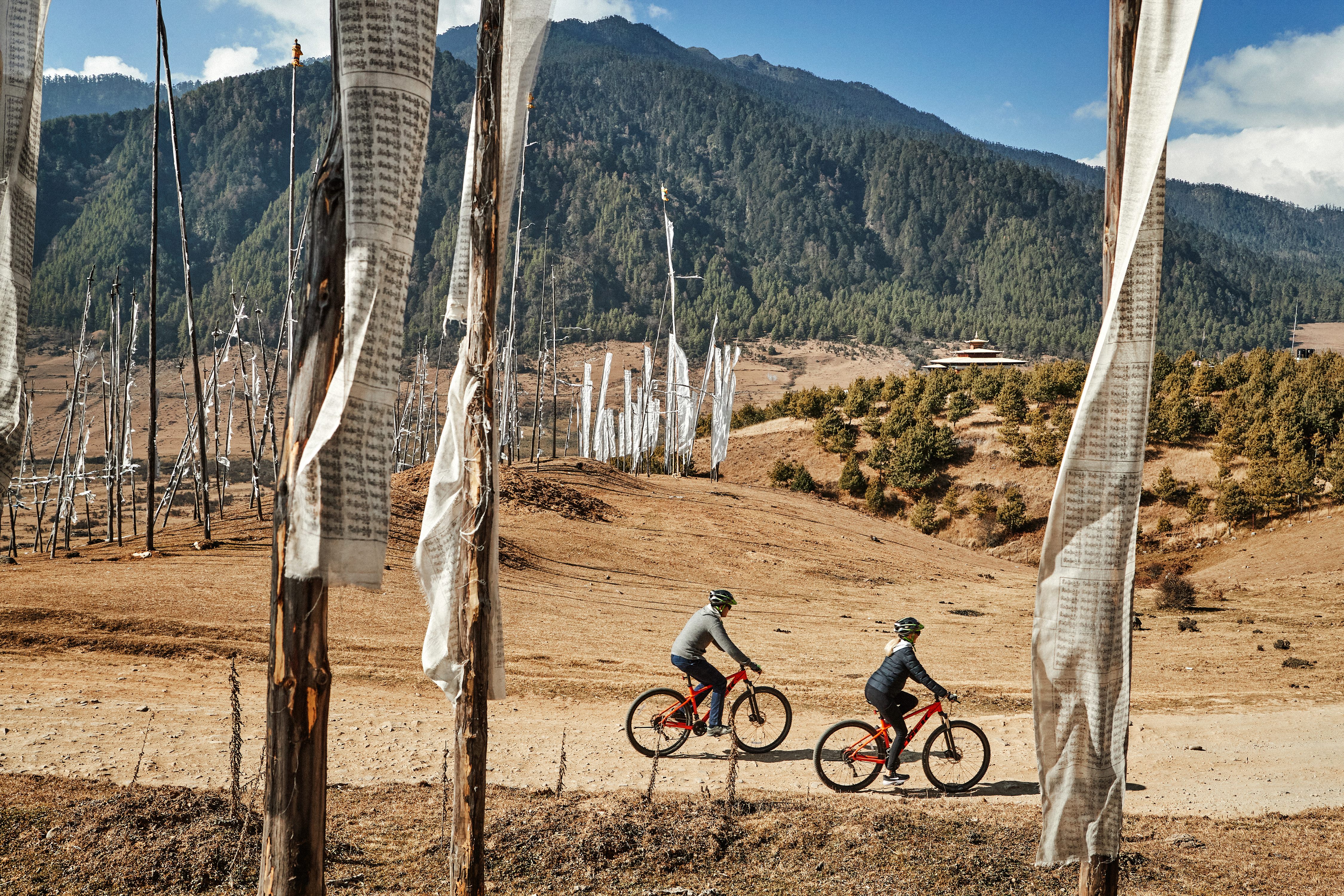 Two cyclists ride along a dirt track beneath tall prayer flags, with brown hills and distant mountains behind them.