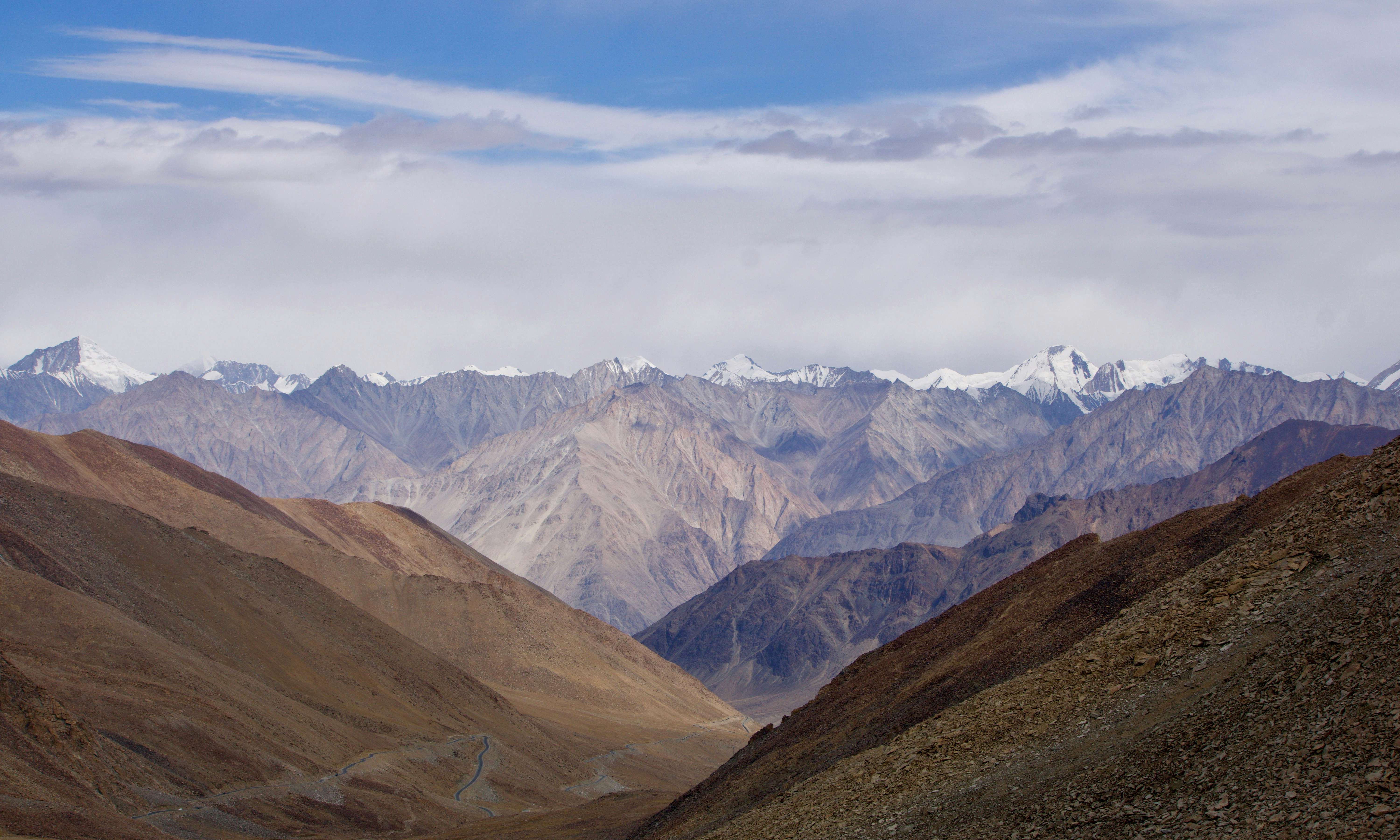 A winding road cuts through a wide mountain valley of brown ridges, with distant snow peaks under blue sky.