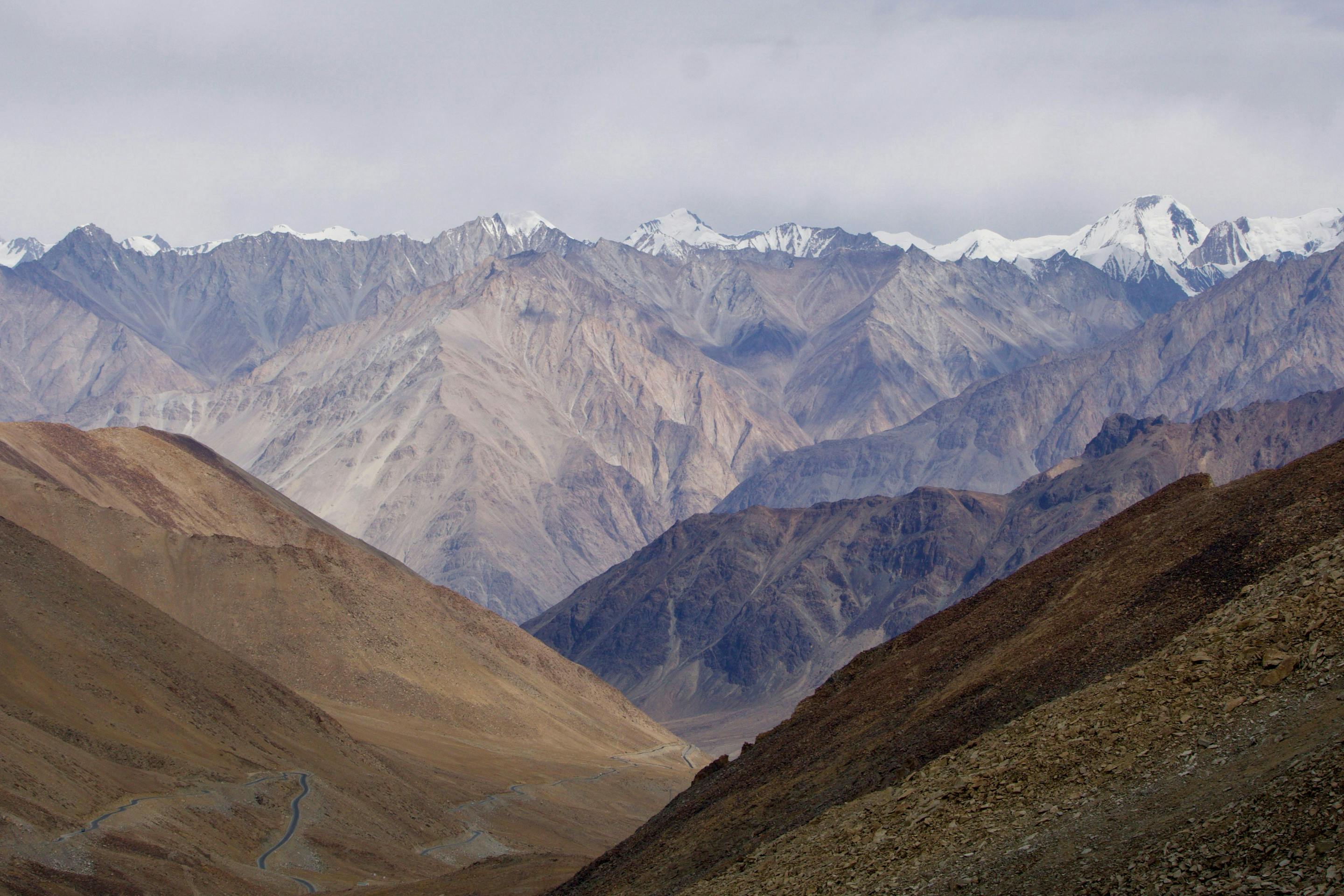 A winding road cuts through a wide mountain valley of brown ridges, with distant snow peaks under blue sky.