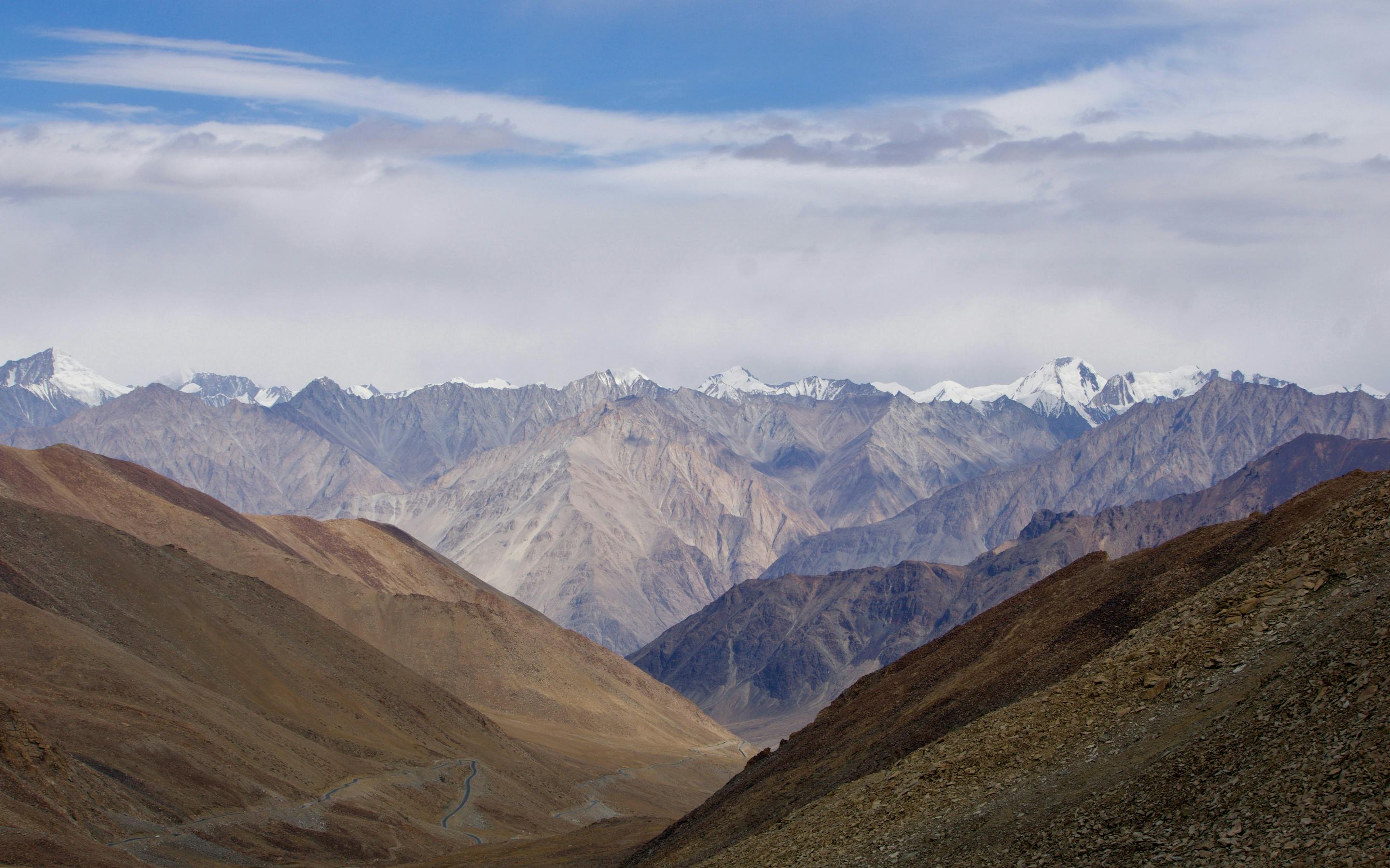 A winding road cuts through a wide mountain valley of brown ridges, with distant snow peaks under blue sky.