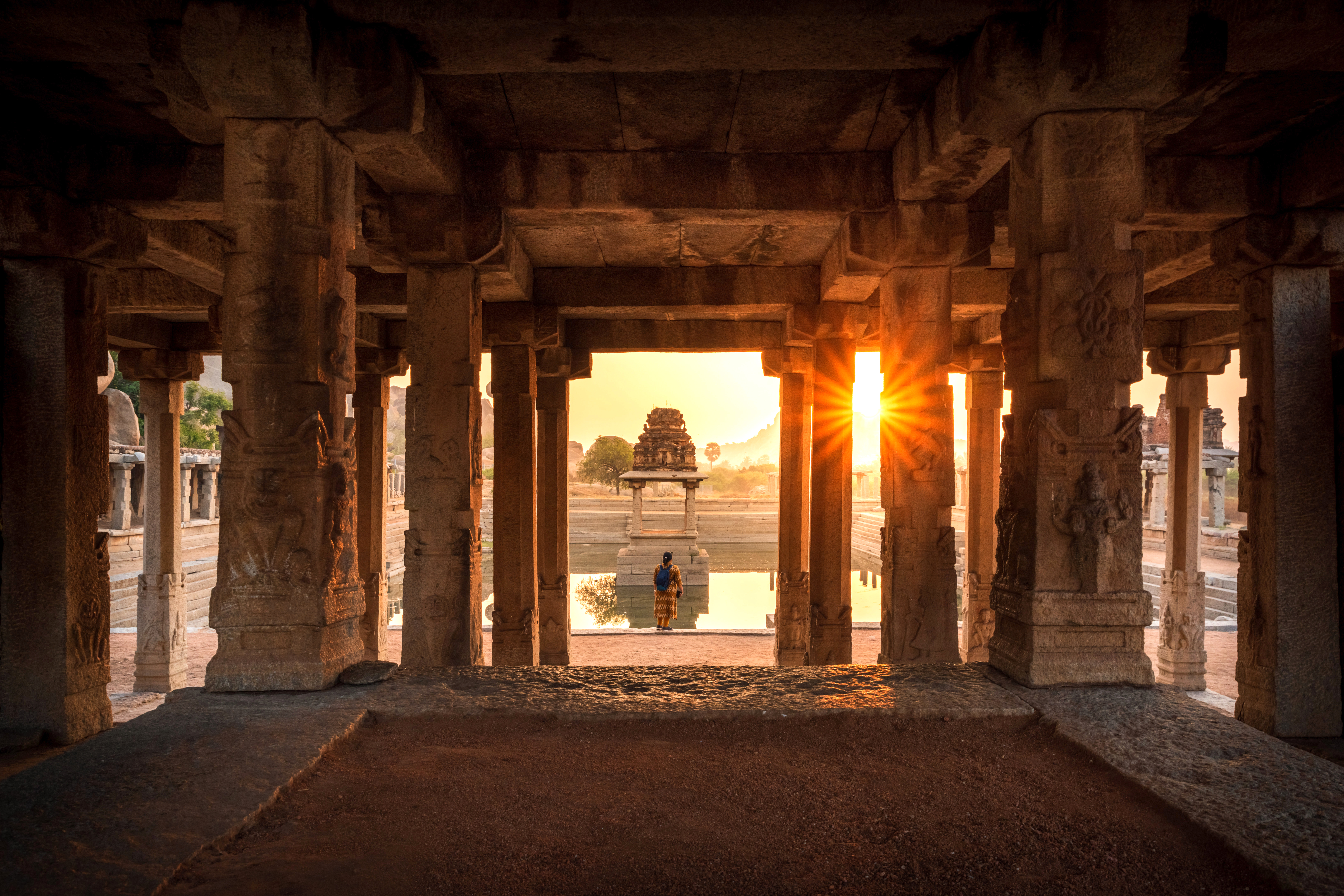 Sunlight streams through a stone-pillared temple corridor, casting long shadows toward a distant shrine at dusk.