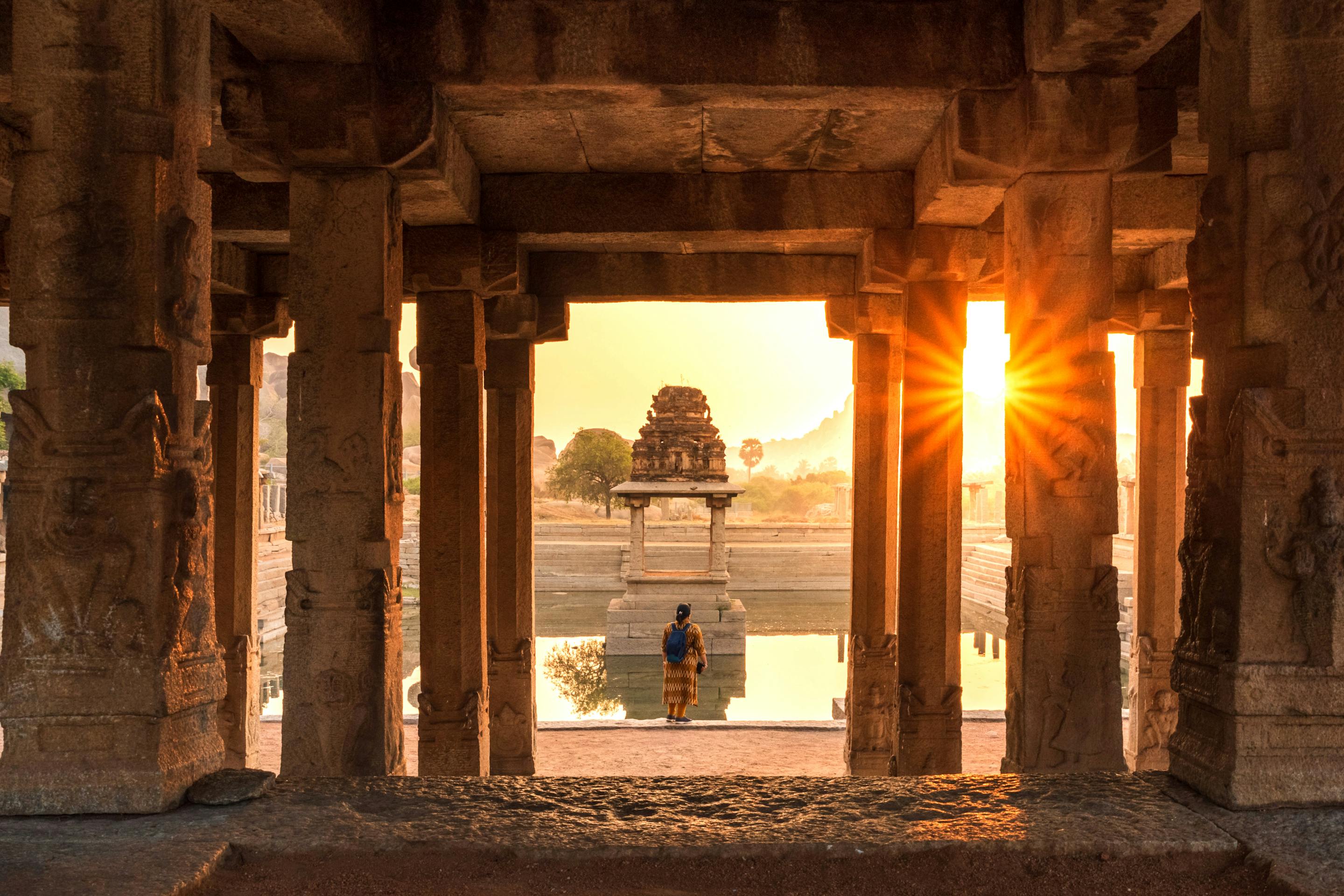 Sunlight streams through a stone-pillared temple corridor, casting long shadows toward a distant shrine at dusk.