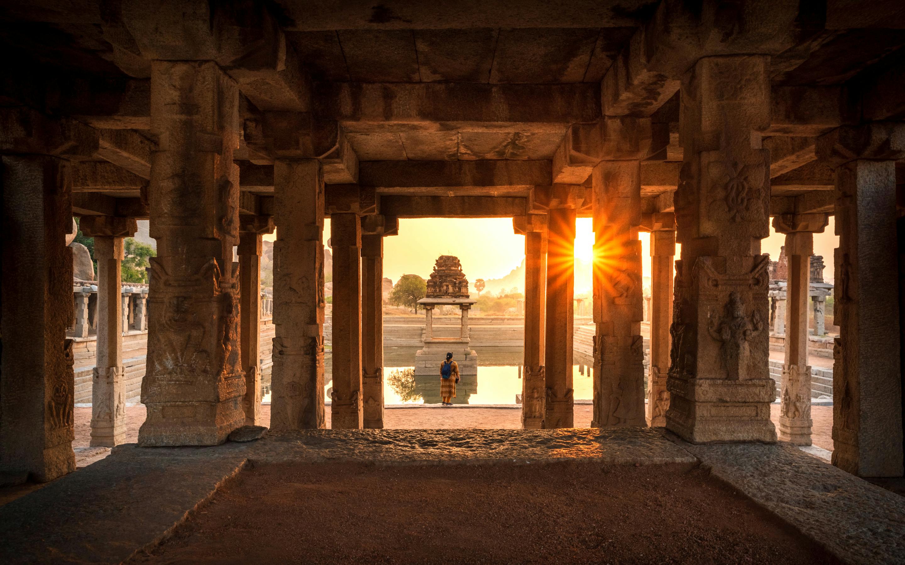 Sunlight streams through a stone-pillared temple corridor, casting long shadows toward a distant shrine at dusk.