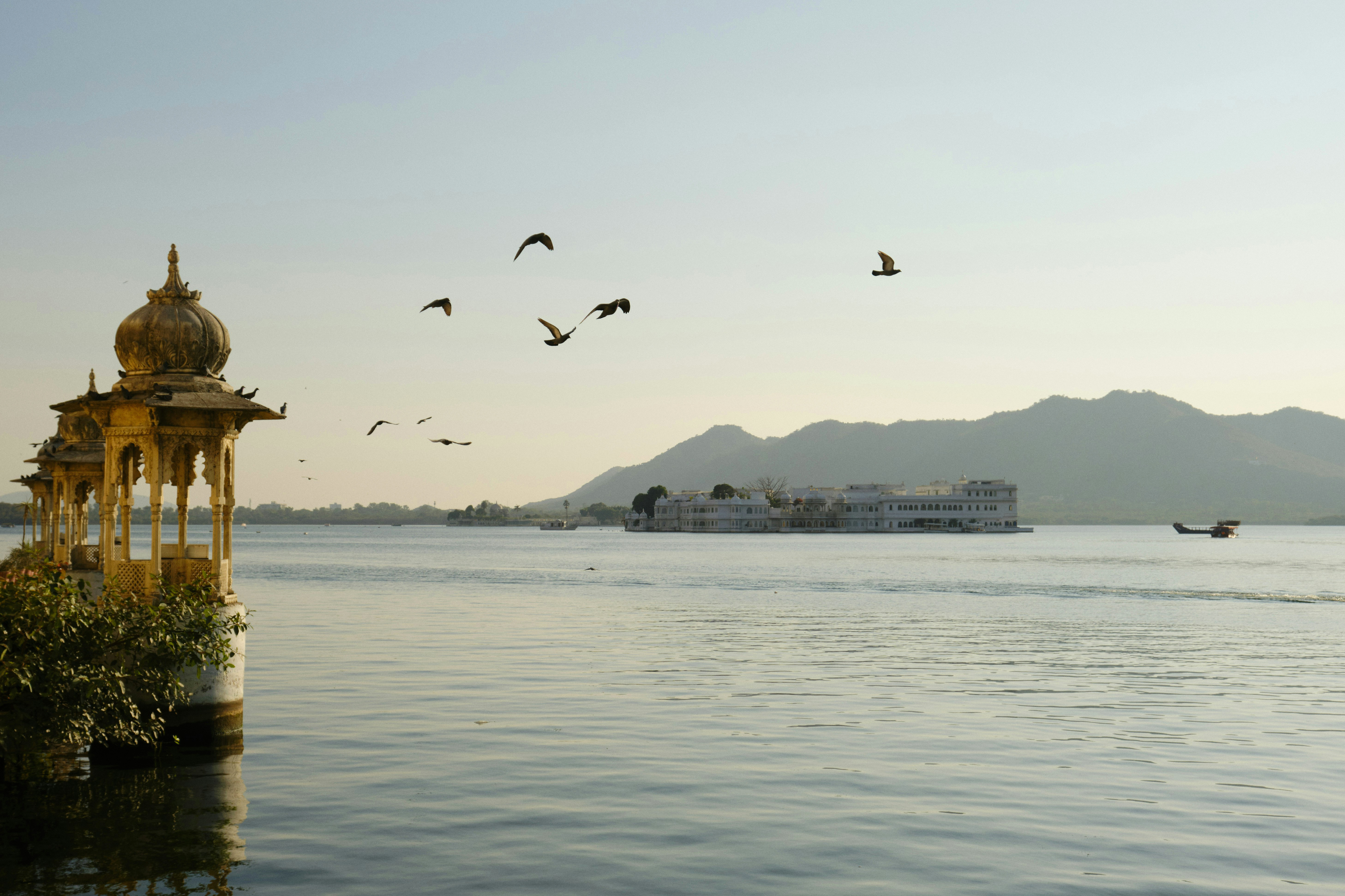 A lakeside pavilion stands near calm water as birds fly overhead, with low mountains and boats in evening light.