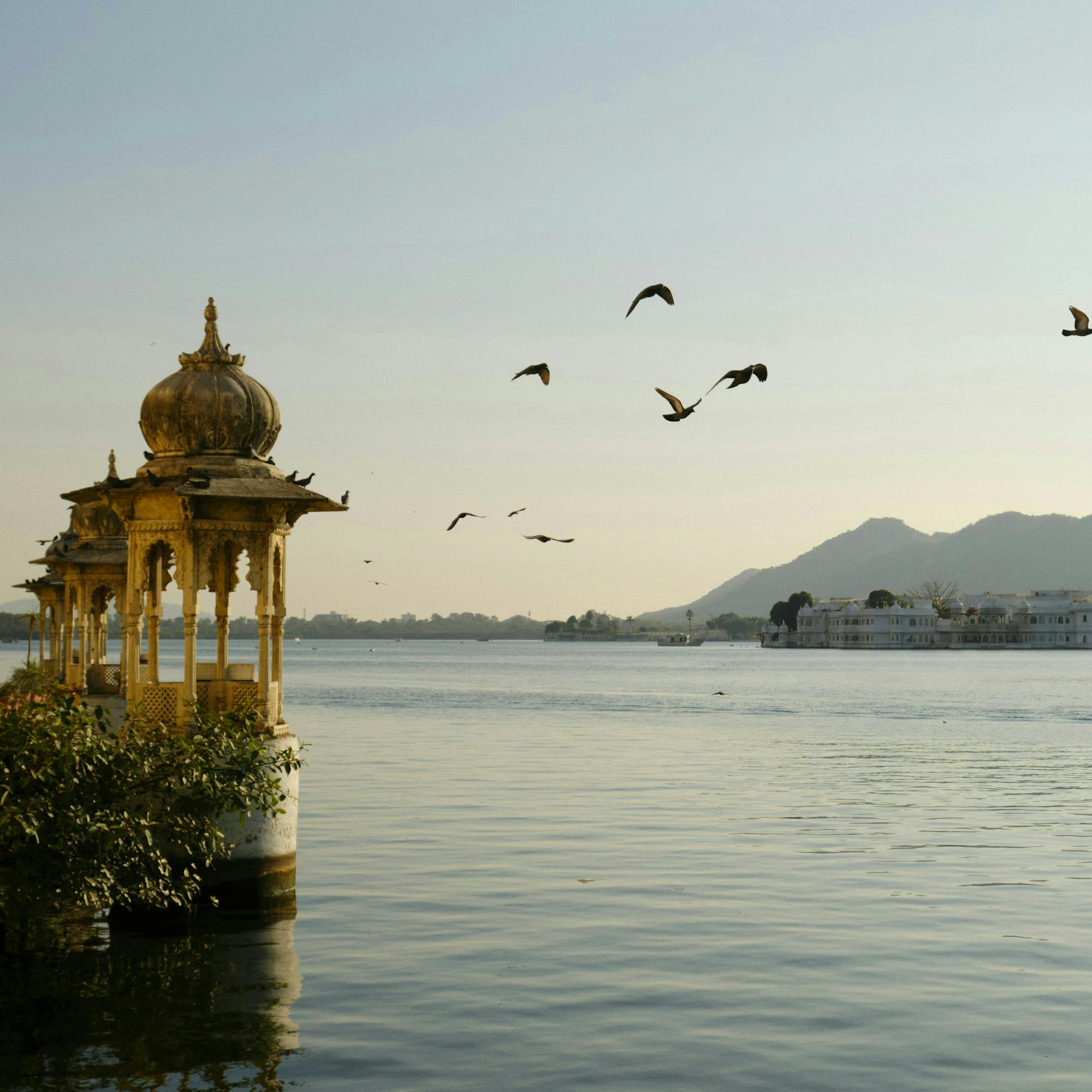 A lakeside pavilion stands near calm water as birds fly overhead, with low mountains and boats in evening light.