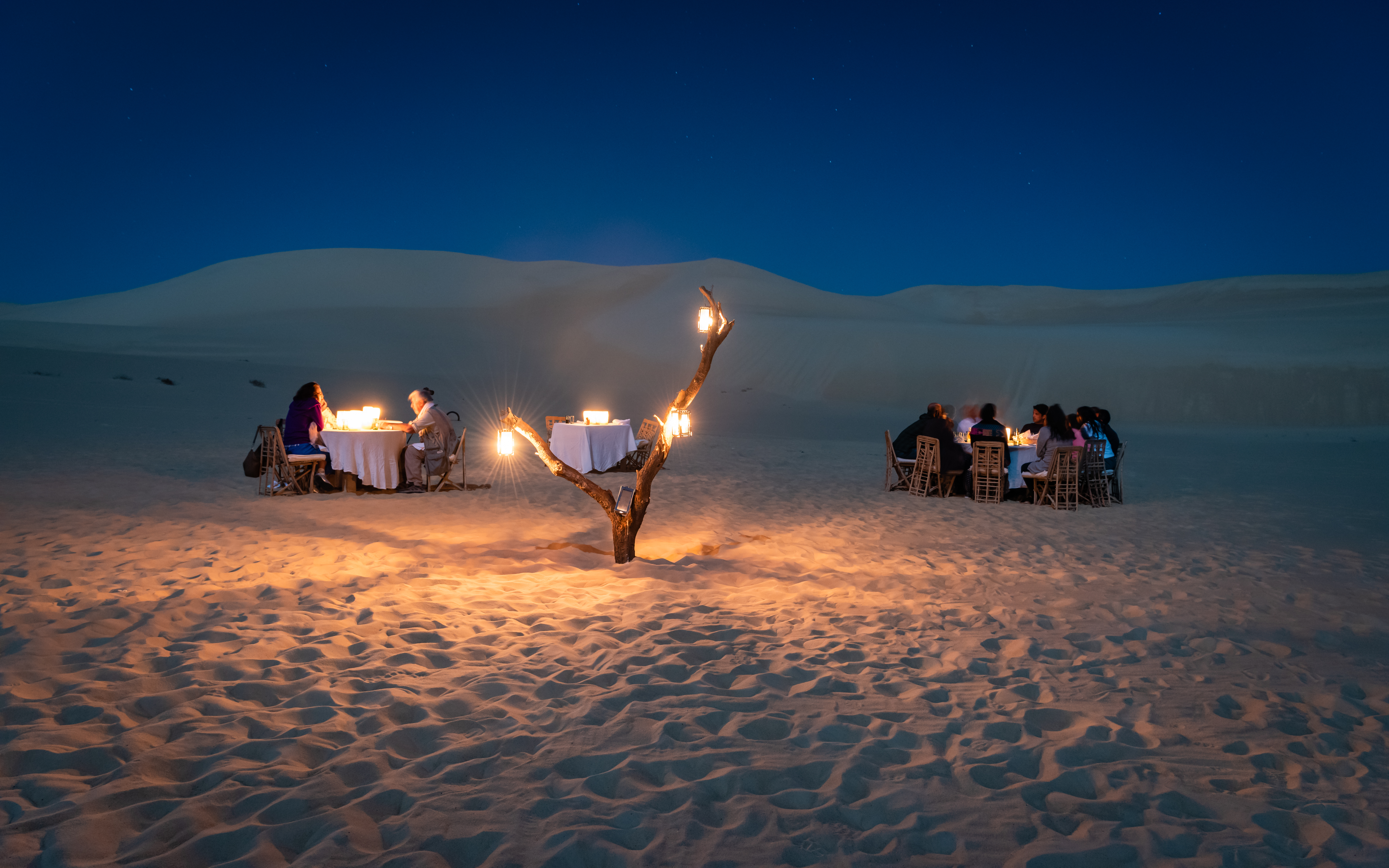 Lantern-lit dinner table set on sand dunes at night, with guests gathered beside a glowing campfire and tents.