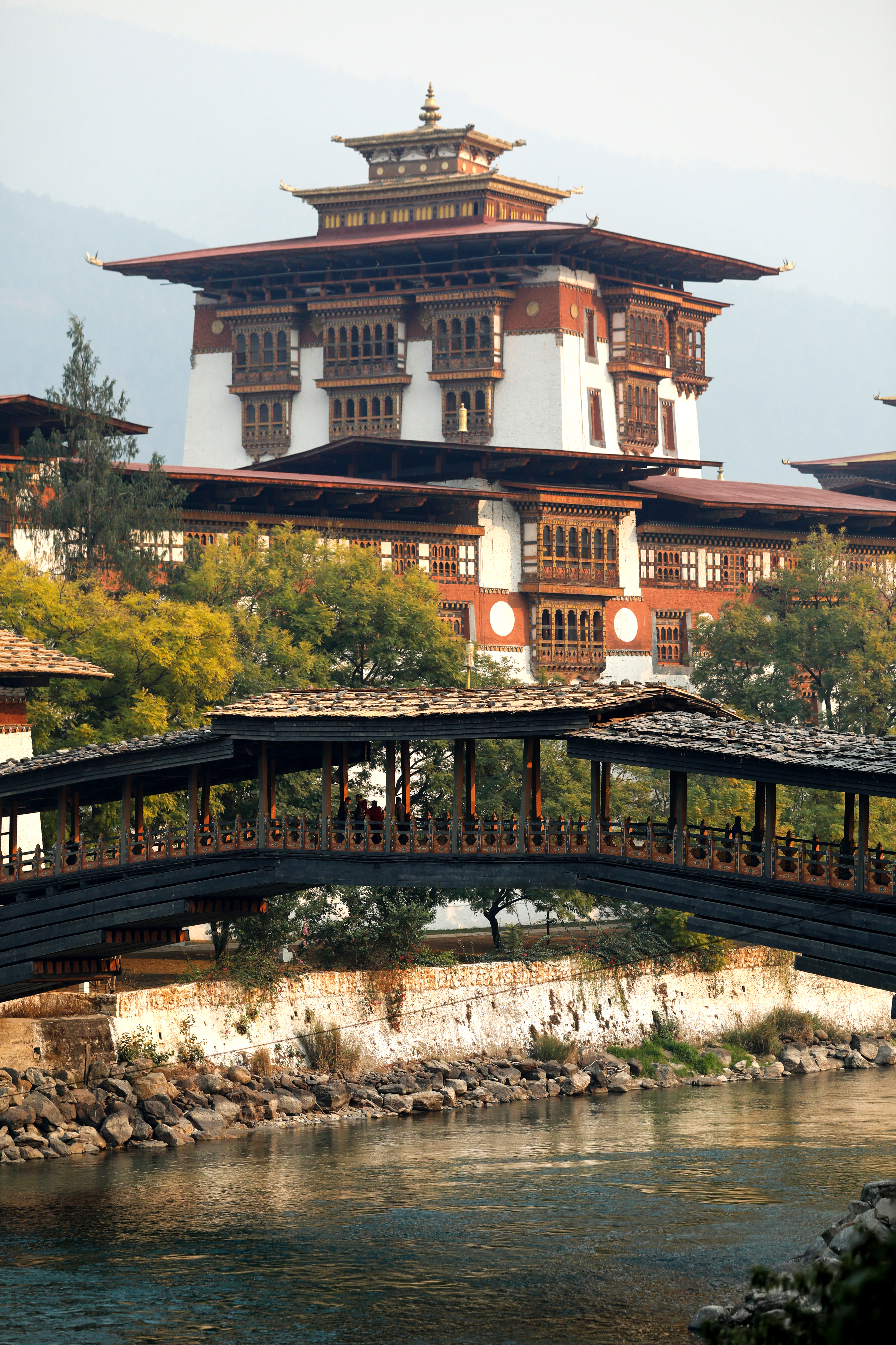 A fortress-like dzong rises beside a river, linked by a wooden bridge and framed by trees and cloudy sky.