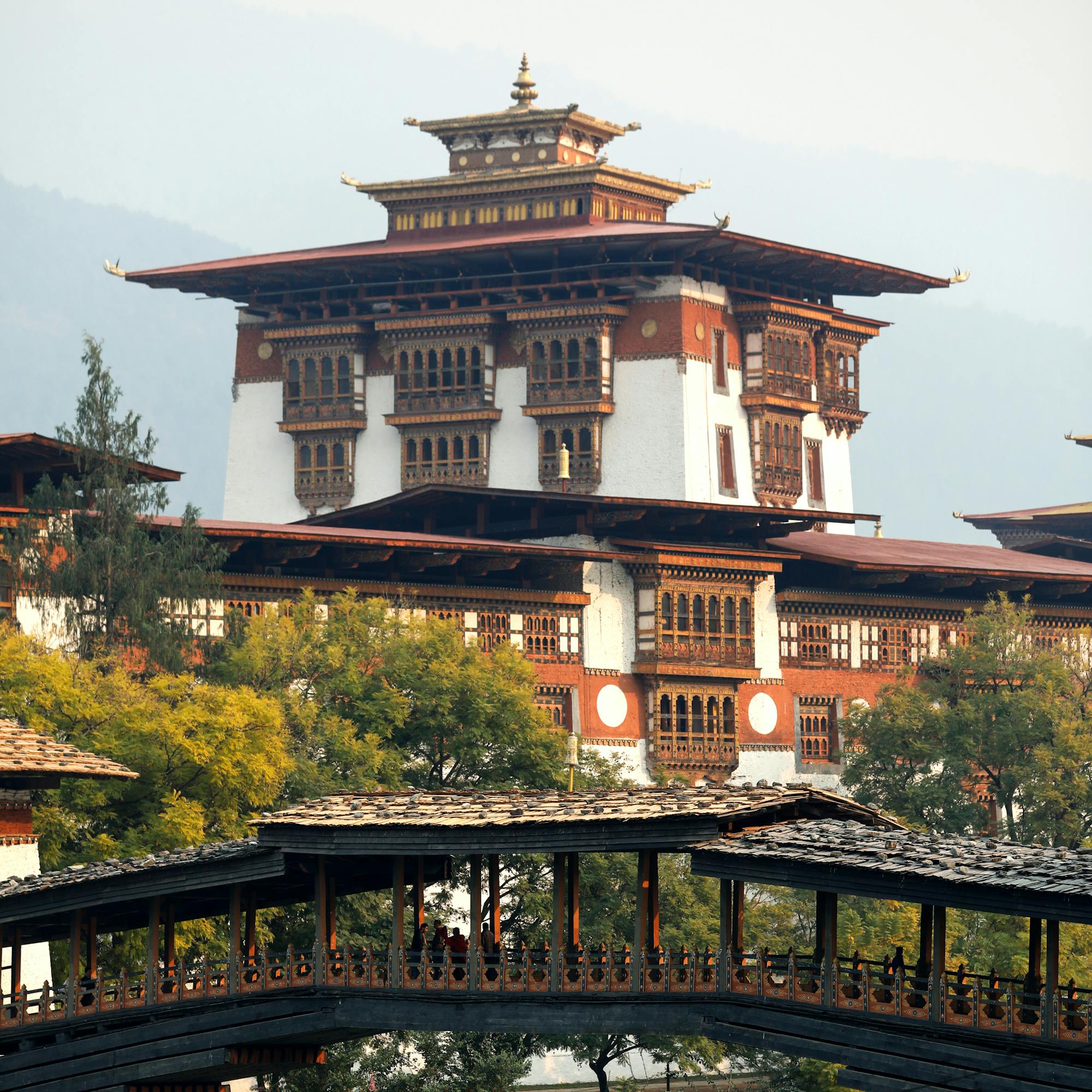 A fortress-like dzong rises beside a river, linked by a wooden bridge and framed by trees and cloudy sky.