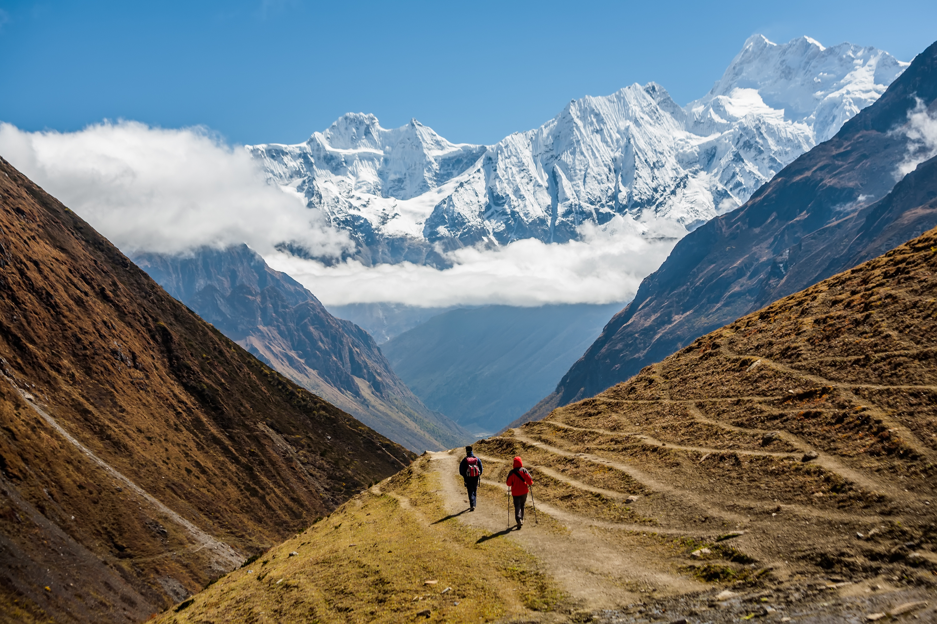 Two hikers follow a winding path through a broad mountain valley, with snowcapped peaks and clouds overhead.