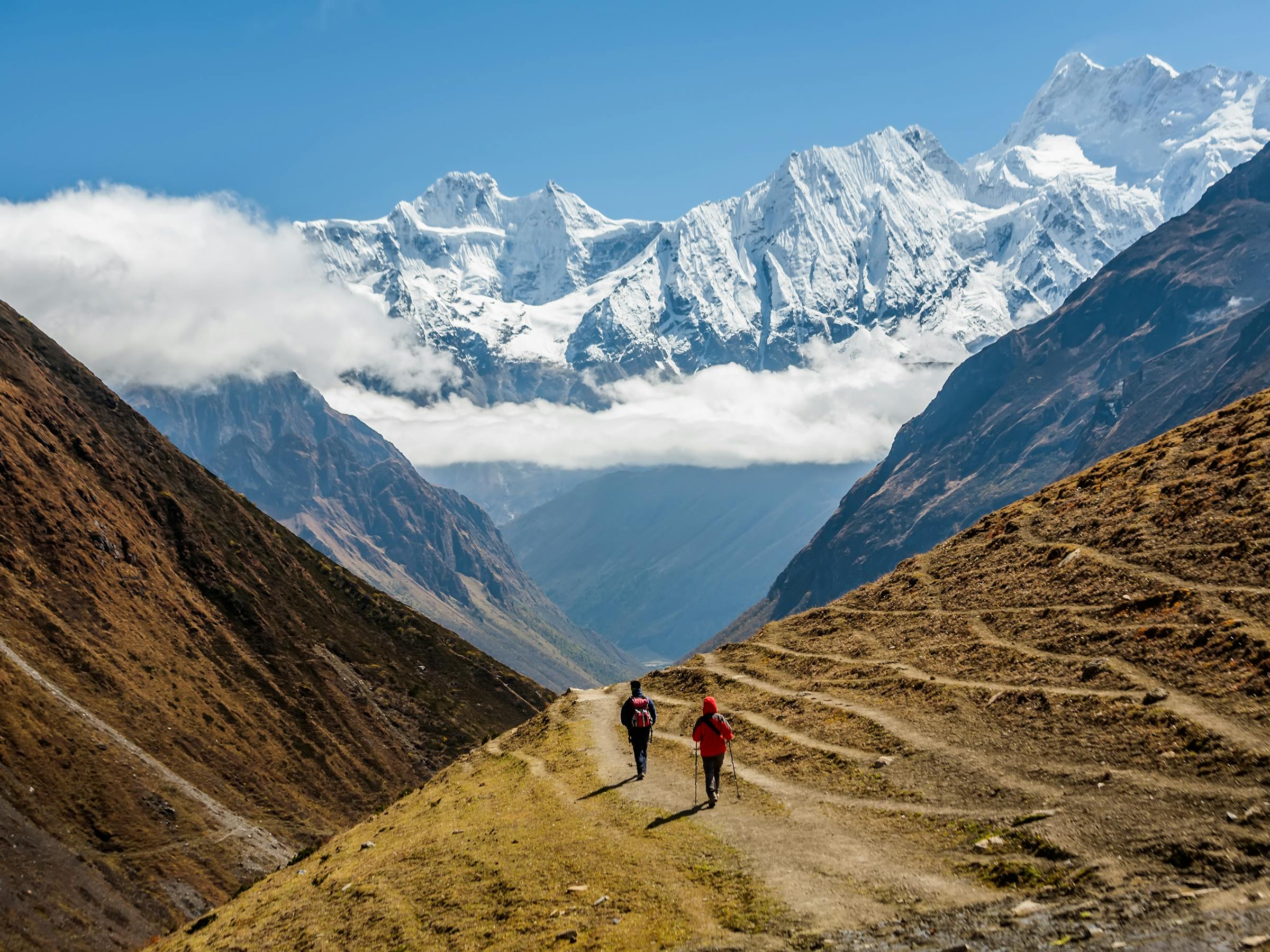 Two hikers follow a winding path through a broad mountain valley, with snowcapped peaks and clouds overhead.