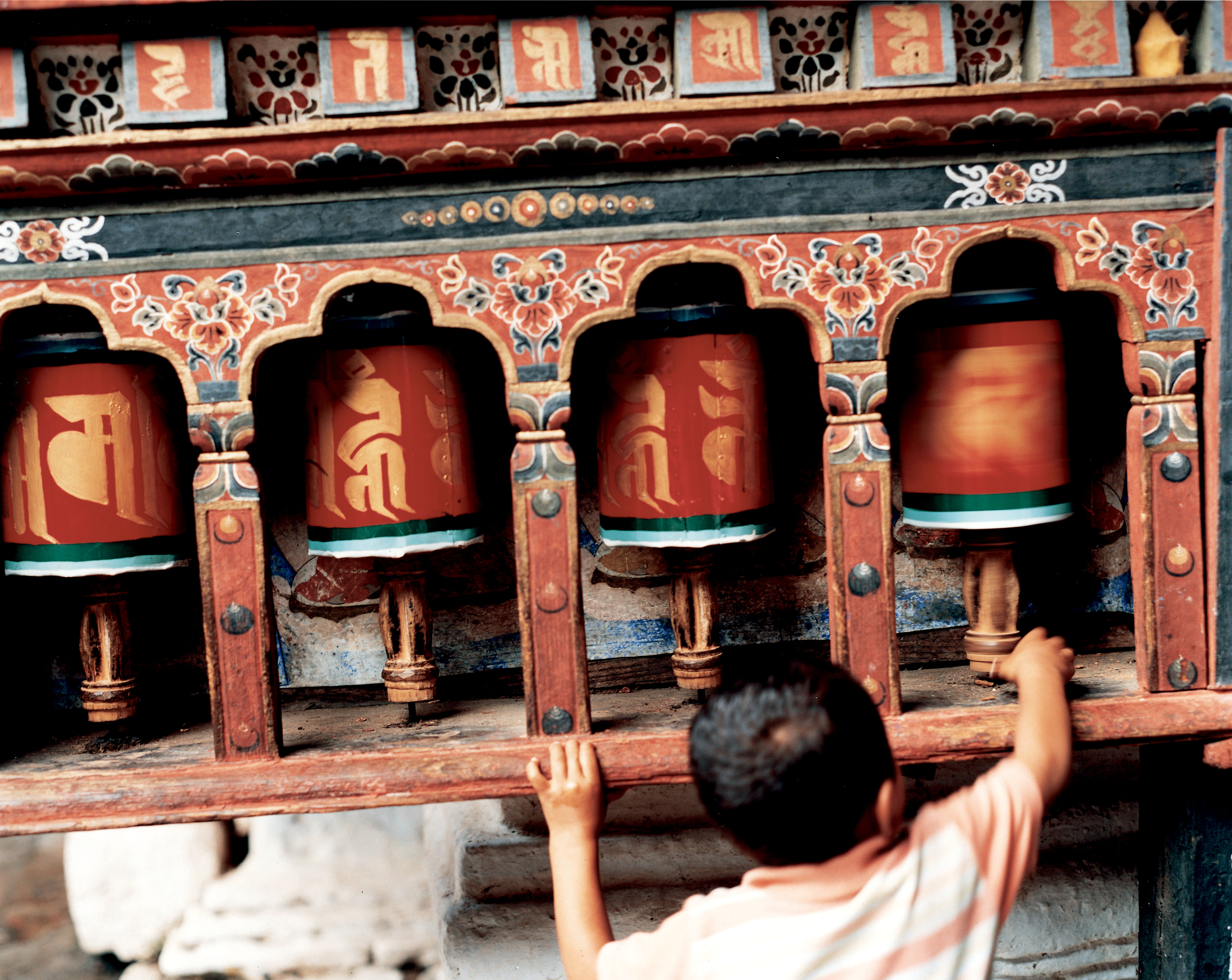 A child reaches up to spin large prayer wheels set into a colorful wall, with carved details and warm sunlight.
