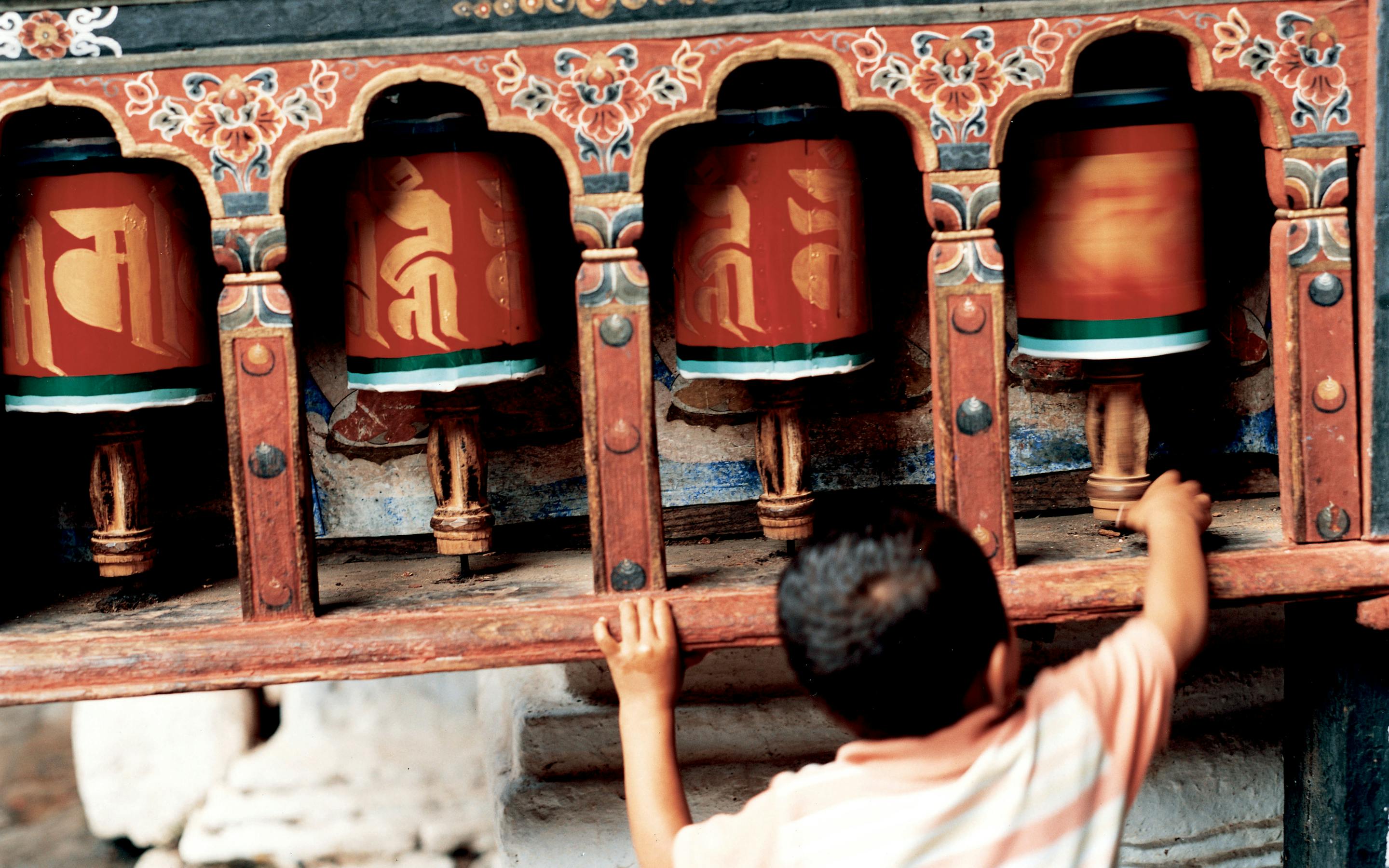 A child reaches up to spin large prayer wheels set into a colorful wall, with carved details and warm sunlight.