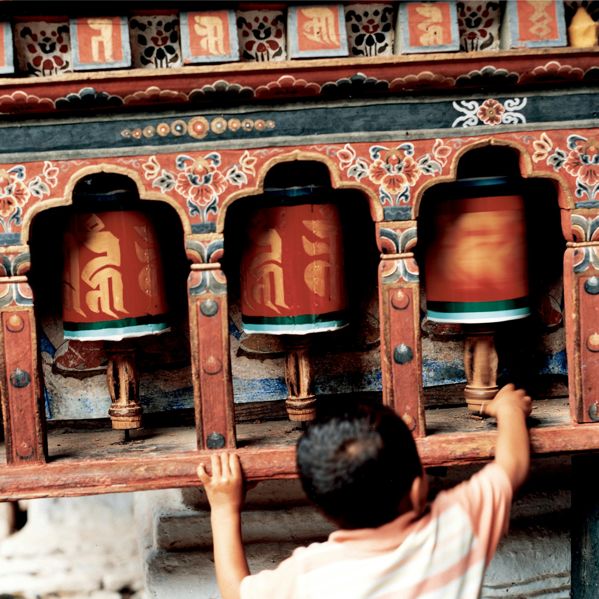 A child reaches up to spin large prayer wheels set into a colorful wall, with carved details and warm sunlight.