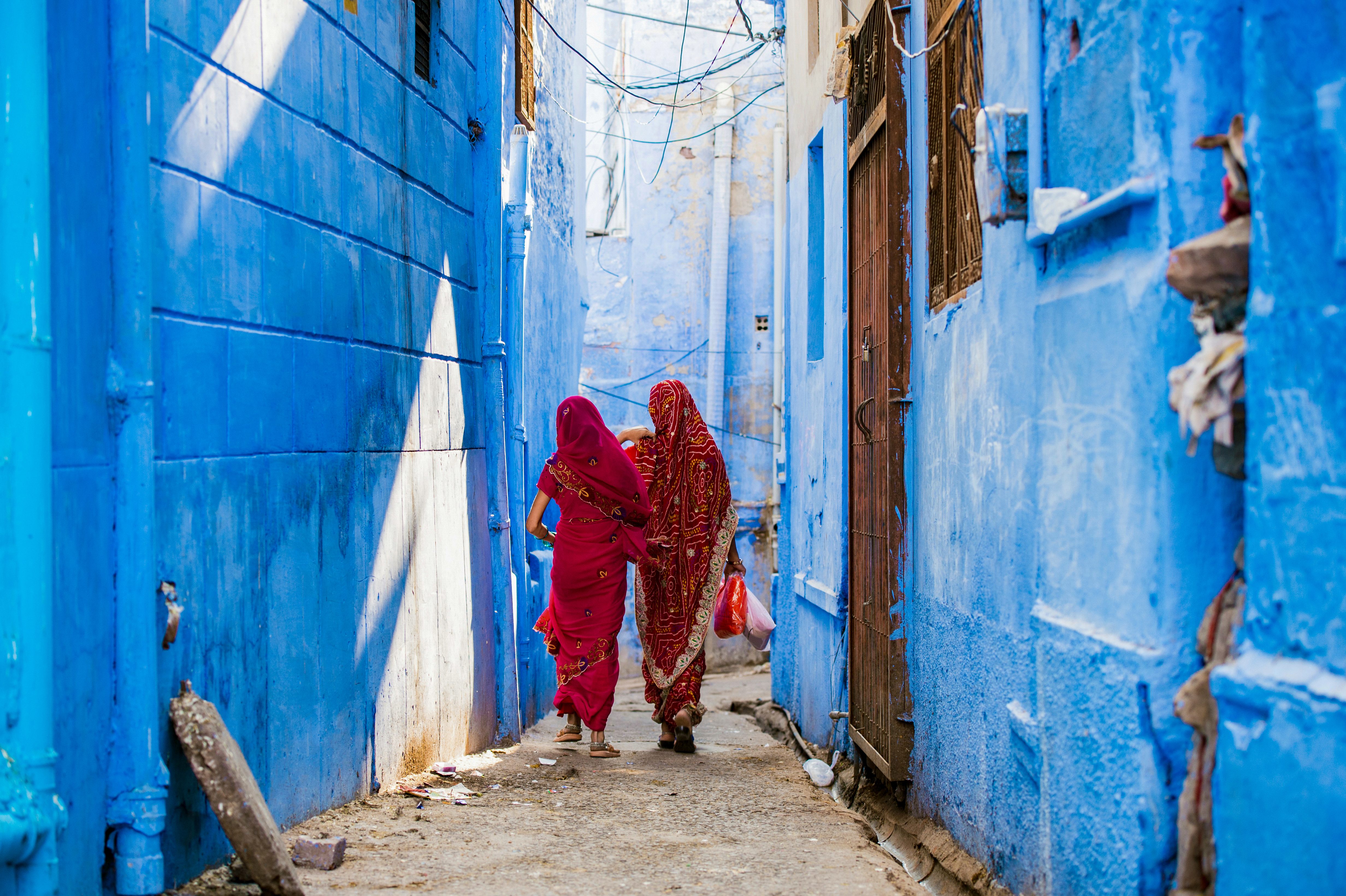 Two women in red saris walk down a narrow blue alleyway, with sunlit walls, doorways, and steps around them.