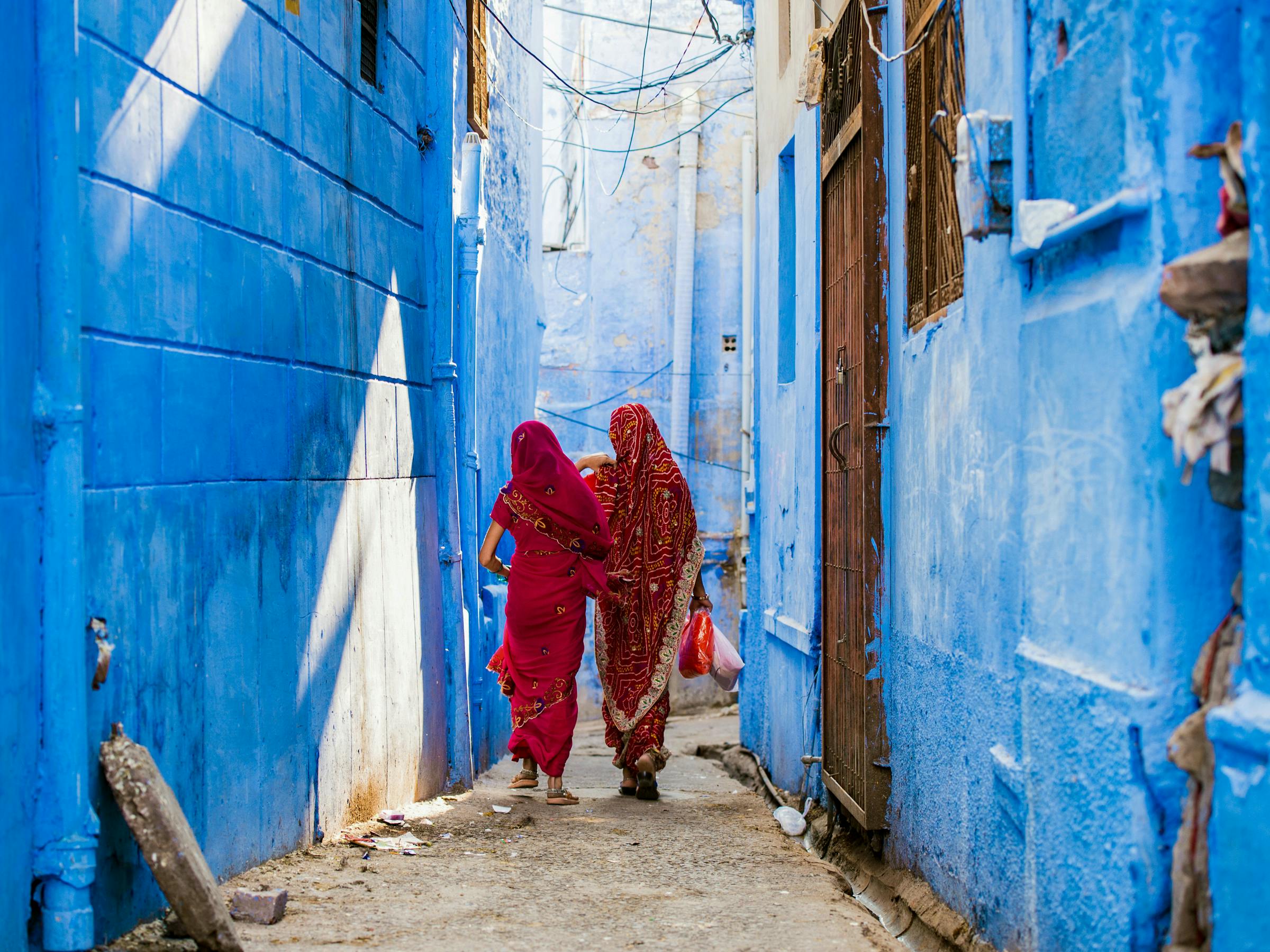 Two women in red saris walk down a narrow blue alleyway, with sunlit walls, doorways, and steps around them.