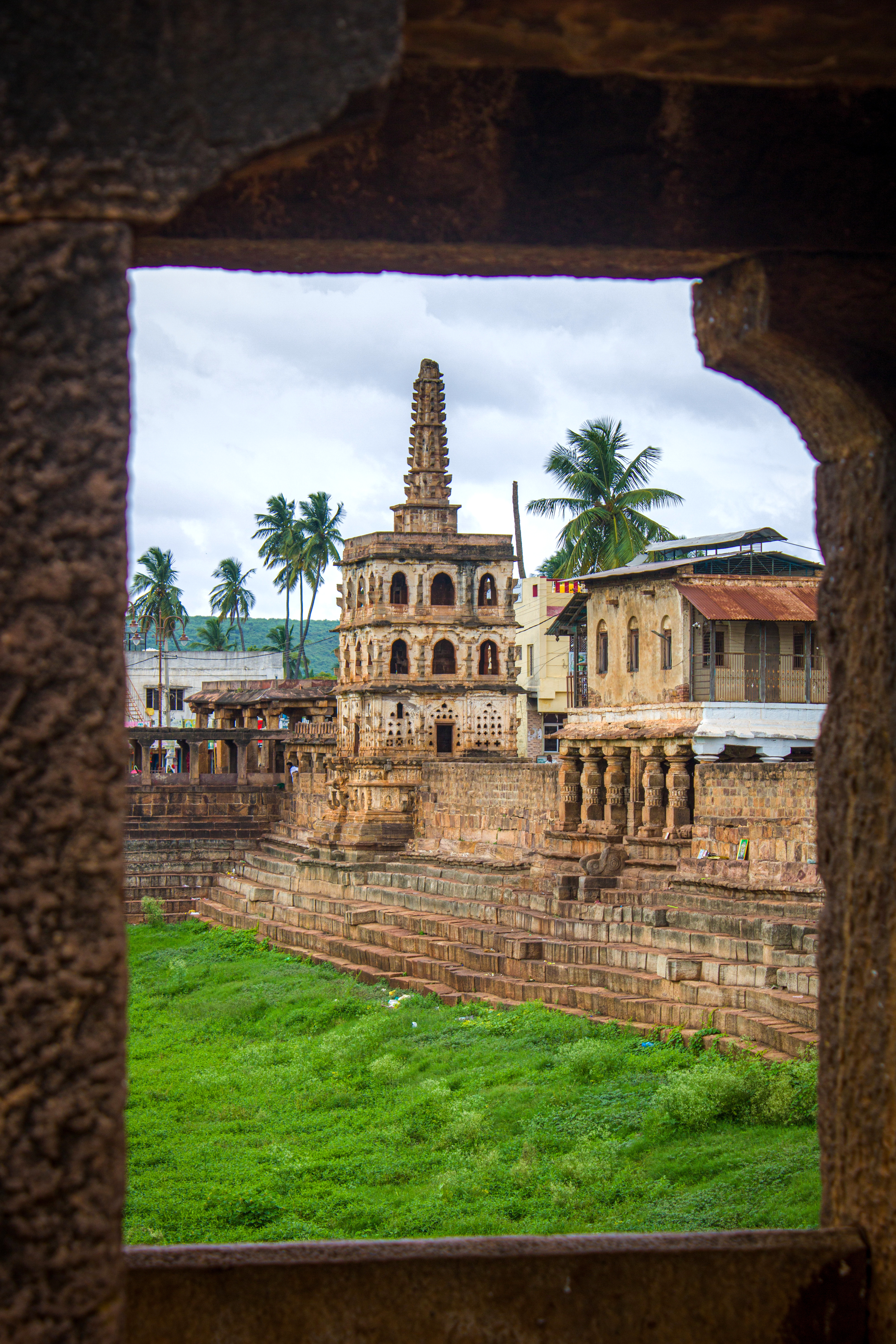 Ancient stone ruins and a small tower are framed by a dark archway, with green grass and palm trees beyond.