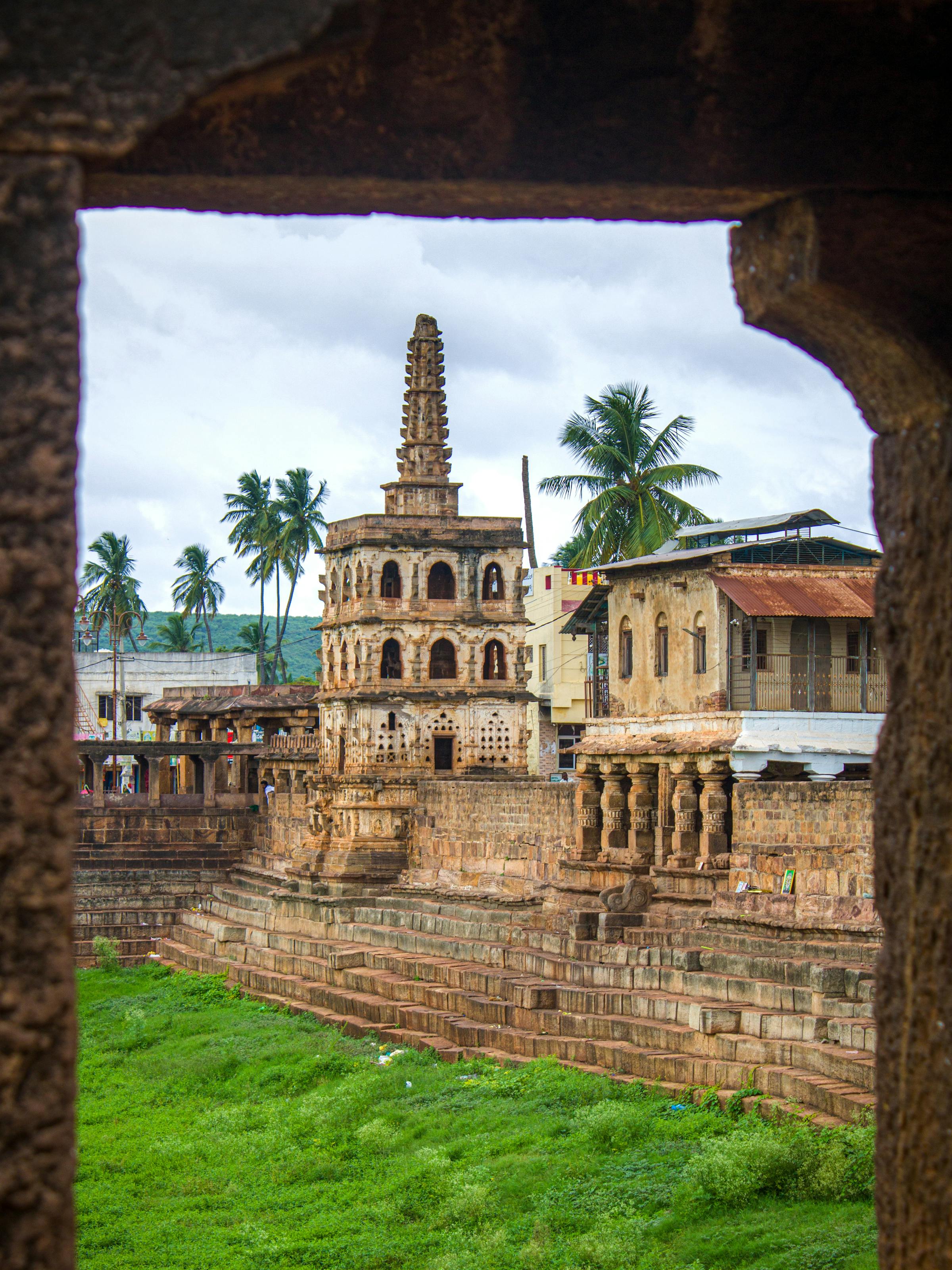 Ancient stone ruins and a small tower are framed by a dark archway, with green grass and palm trees beyond.
